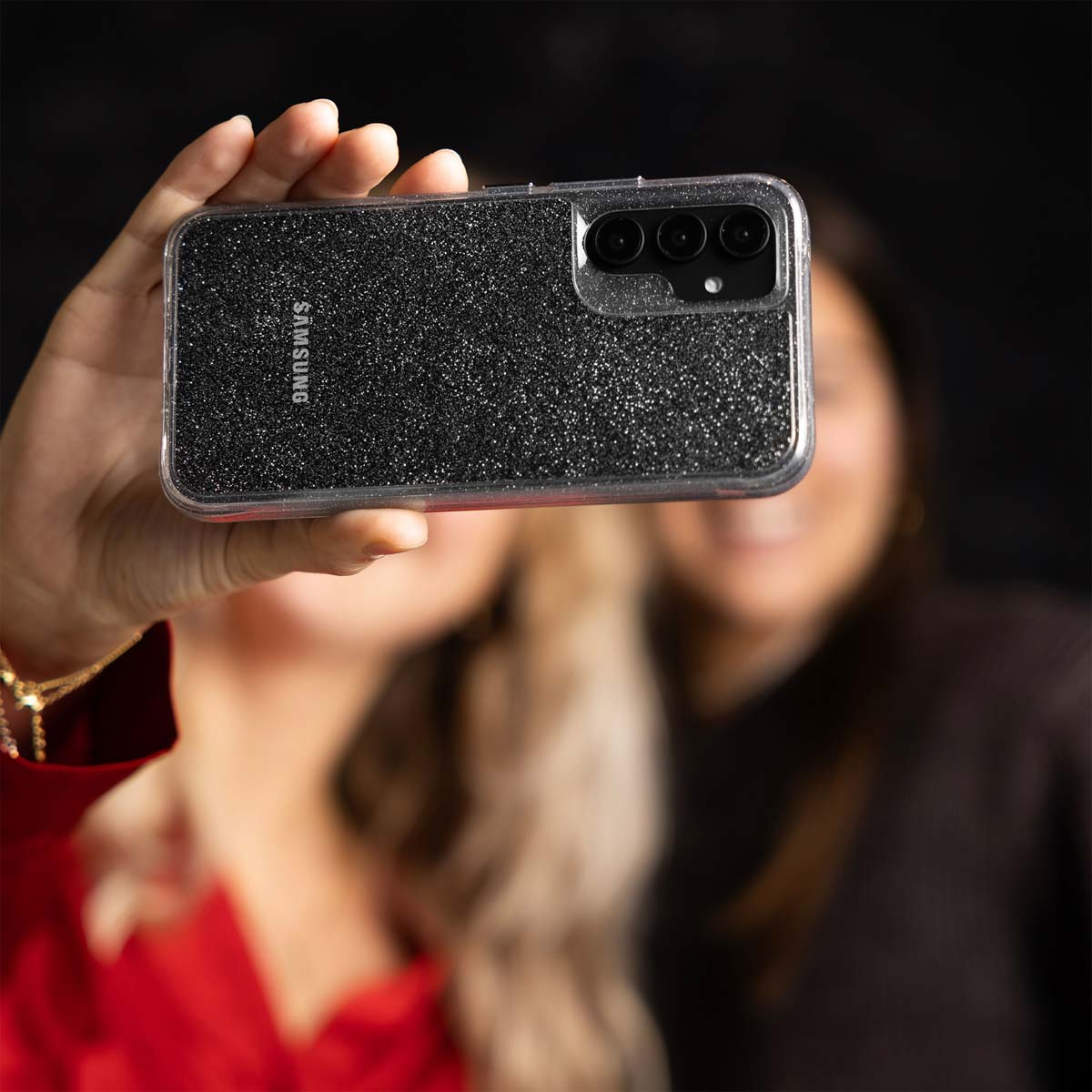 Een persoon houdt een telefoon met een glitterhoesje vast. Twee andere personen op de achtergrond.
