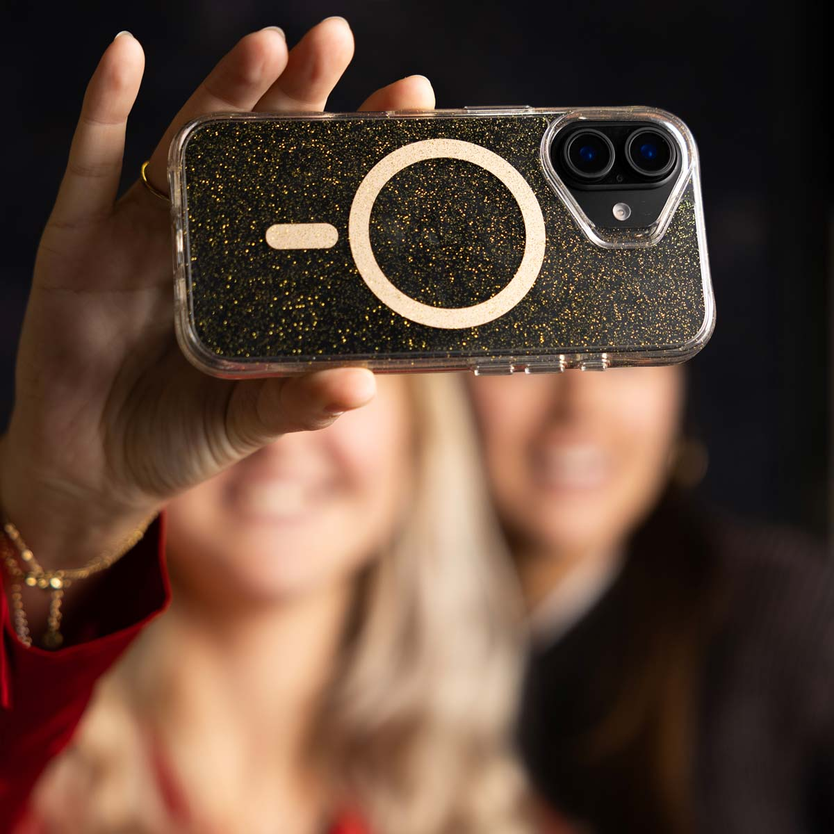 Une main tenant une coque de téléphone avec un anneau et une caméra. Deux femmes floues en arrière-plan.