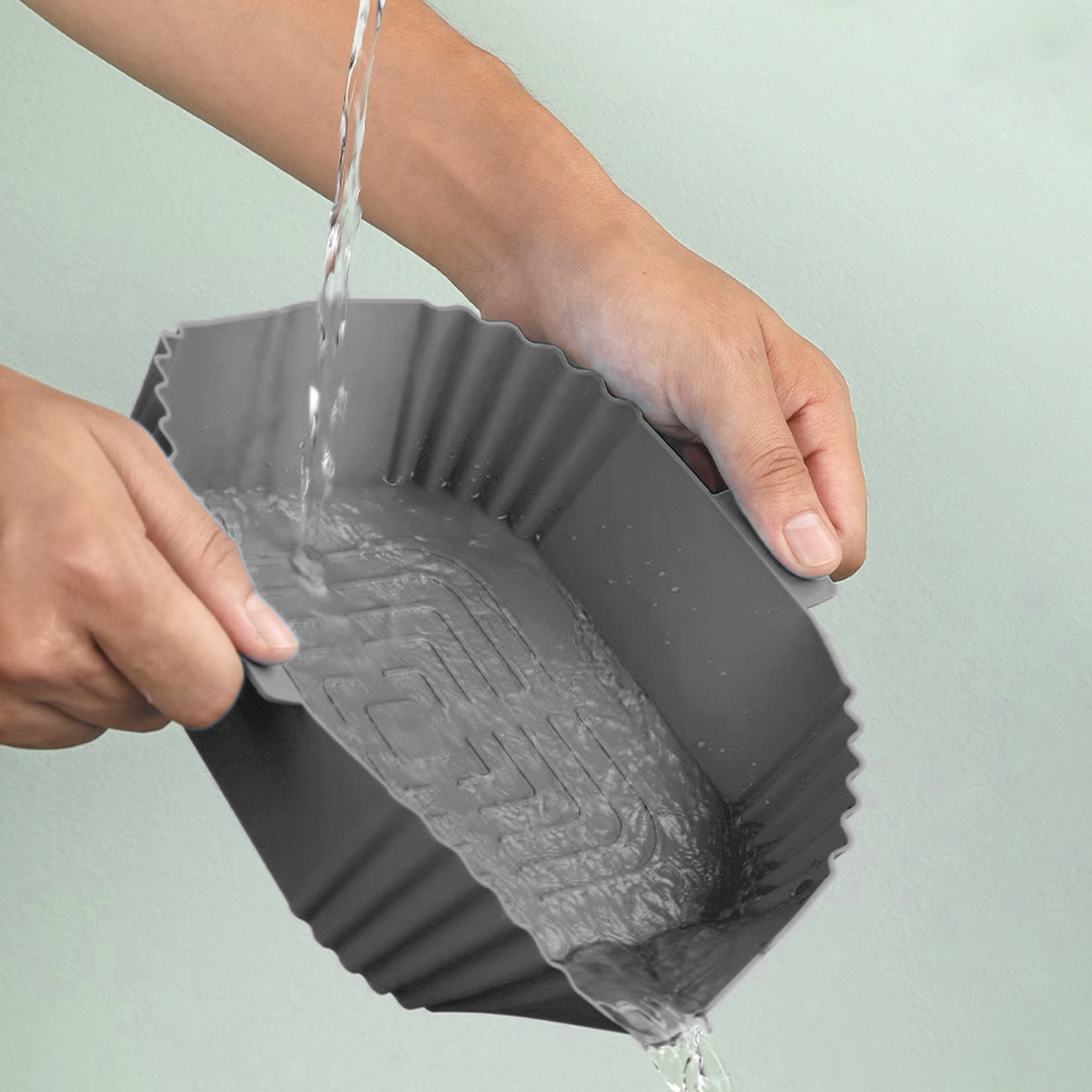 Hände halten einen grauen, geriffelten Silikon-Airfryer-Korb, Wasser gießt hinein.