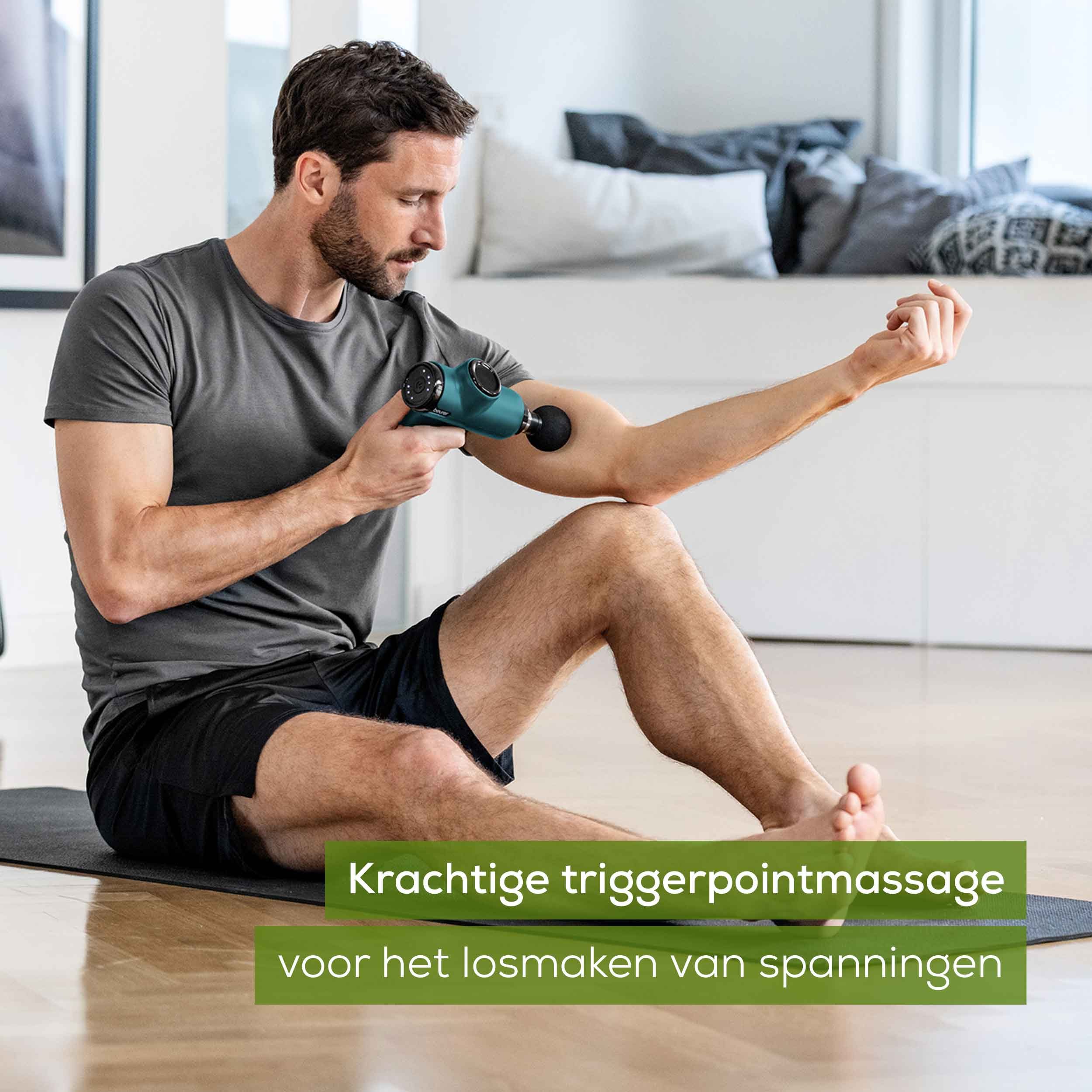 Een bebaarde man in een grijs t-shirt gebruikt een massagepistool op zijn bovenarm.