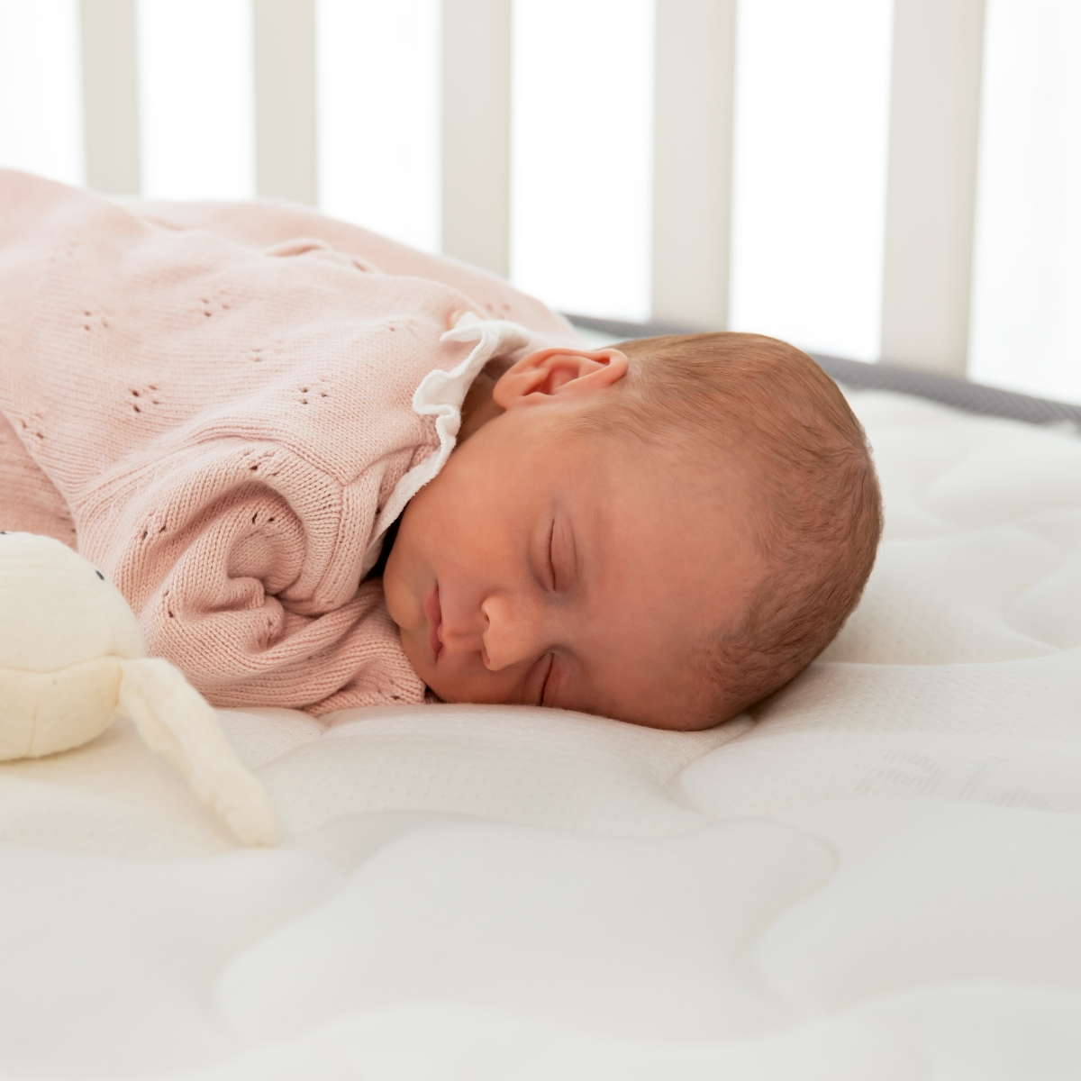 Un bebé durmiendo en un colchón blanco, envuelto en una manta rosa.