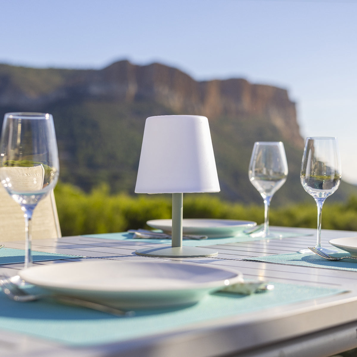 Tischset mit Lampe, Weingläsern und Tellern. Hintergrund zeigt Berg und grüne Bäume unter sonnigem Himmel.