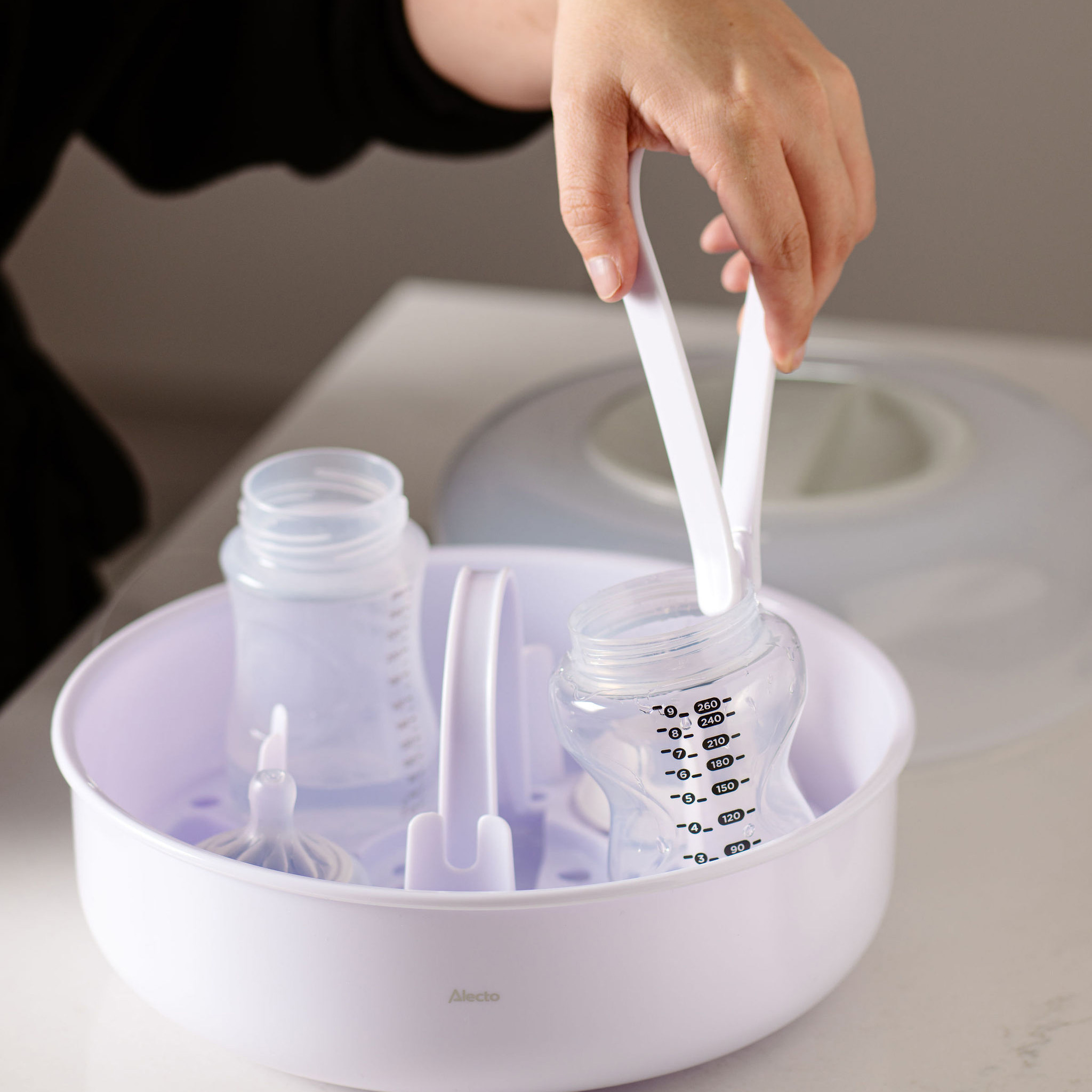 Una mano usando pinzas para sacar un biberón de un esterilizador blanco que contiene otras botellas.