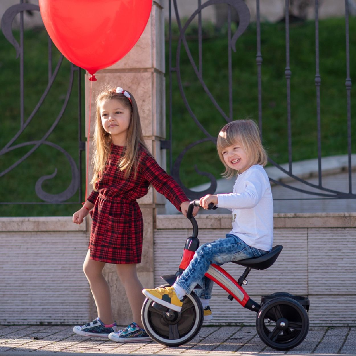 Dos niños con un globo rojo y un triciclo. Fondo verde y luz solar.