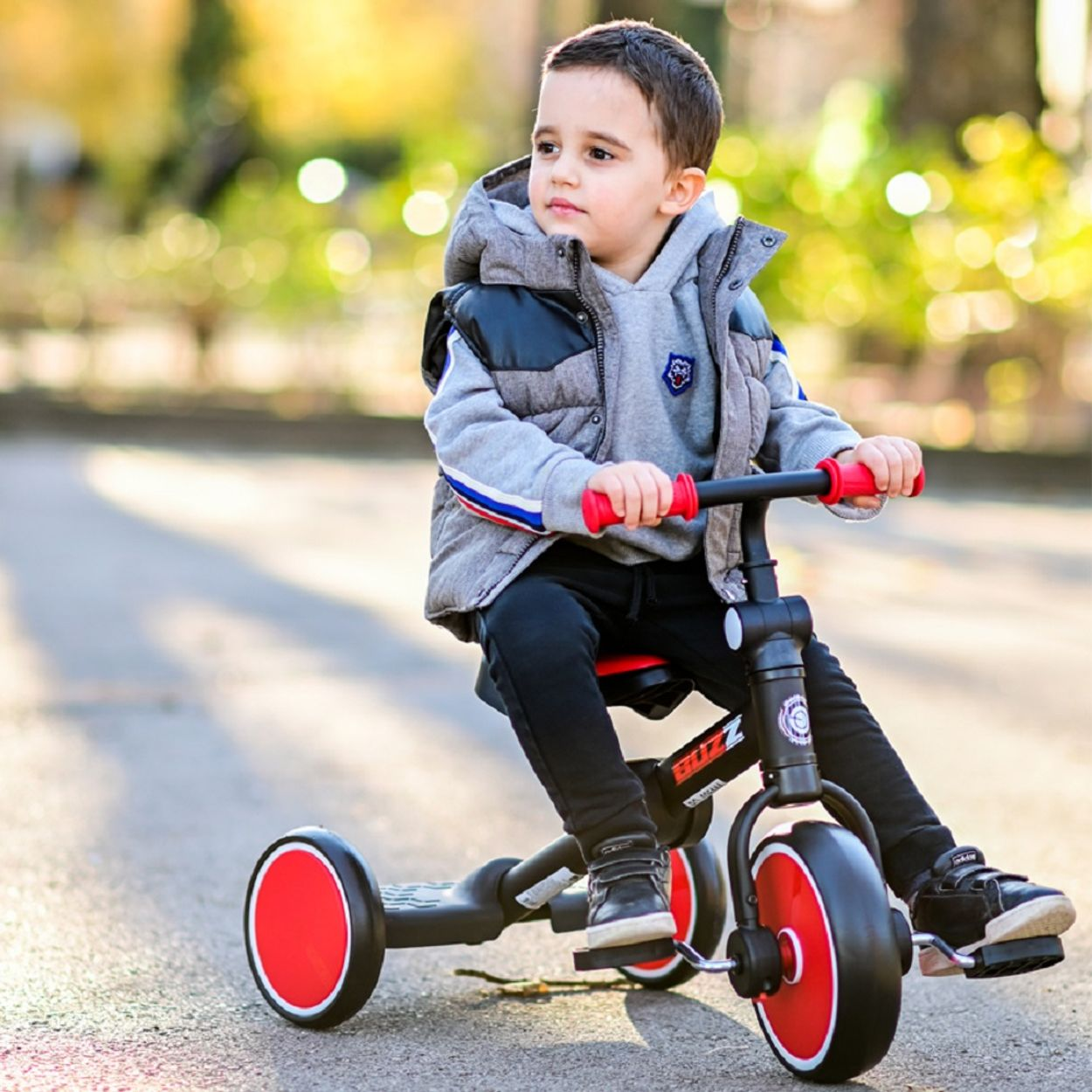 Un niño pequeño monta en un triciclo rojo y negro en un camino pavimentado, fondo soleado.