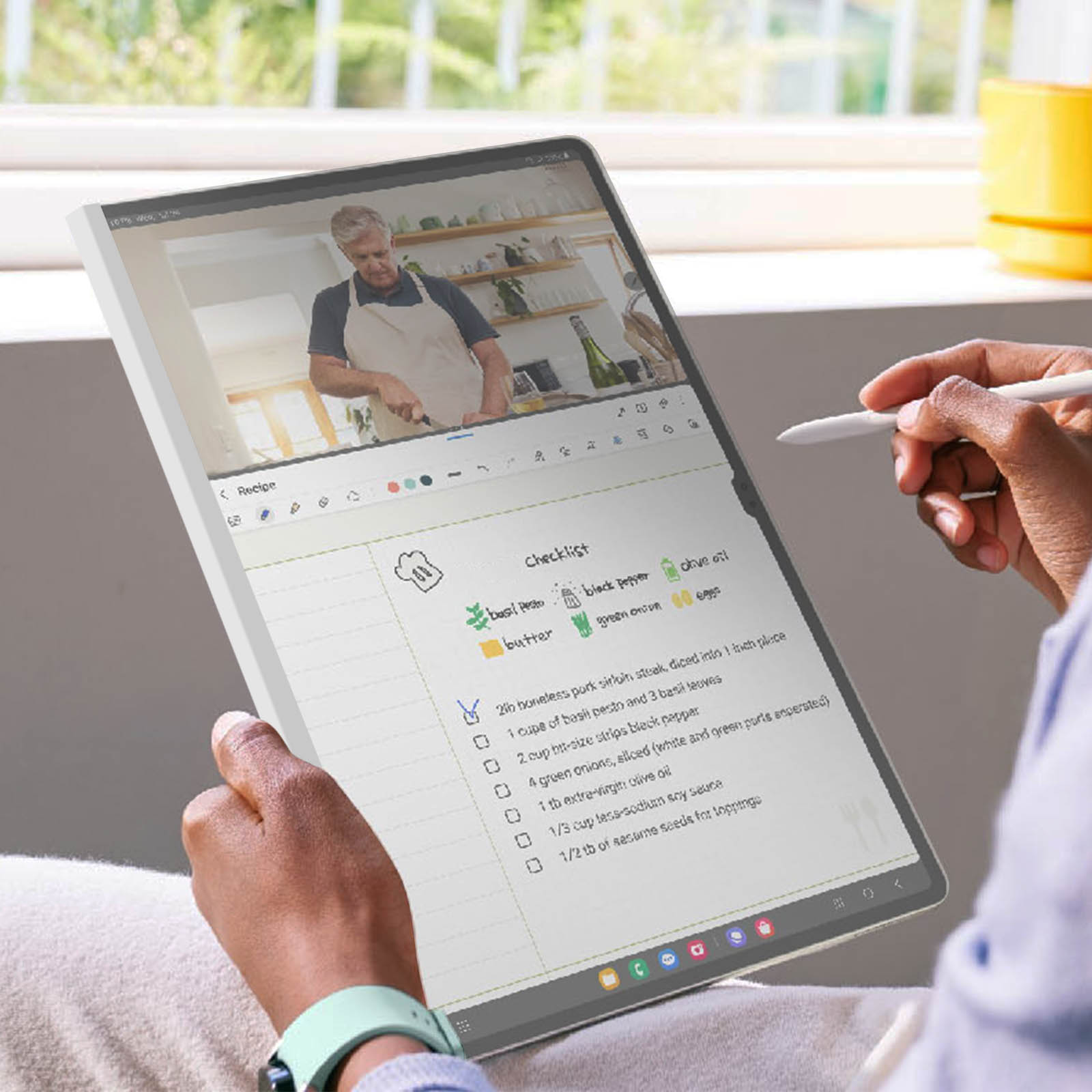 Una persona usa uno stilo su un tablet che mostra una ricetta. Una persona sta cucinando sullo sfondo.