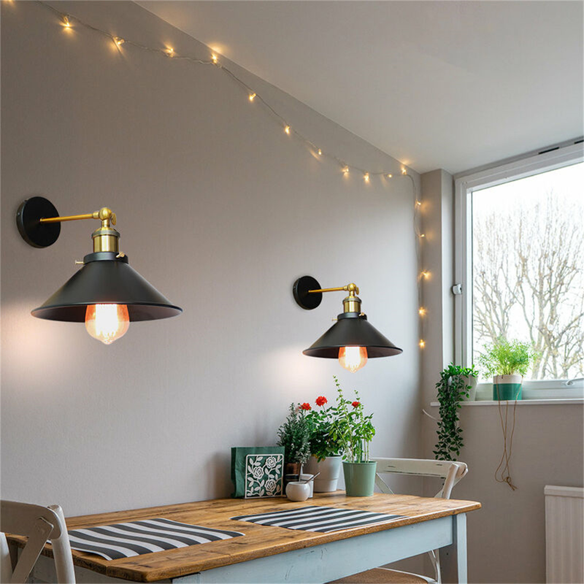 Zwei schwarze Lampen mit goldenen Armen an grauer Wand über Tisch. Lichterkette und Fenster sichtbar.