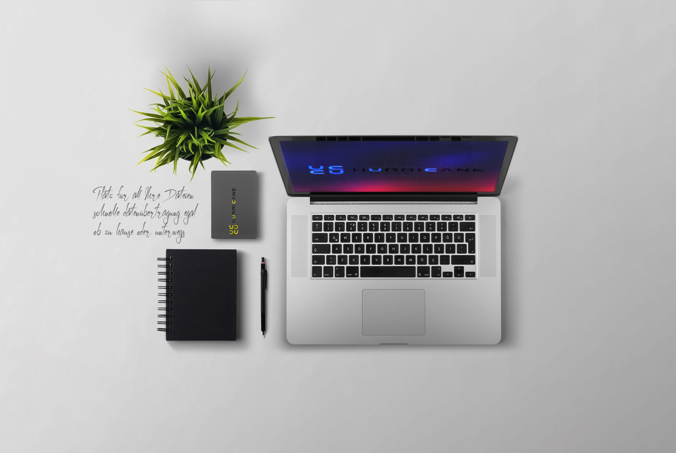 Laptop, Notizbuch, Pflanze und ein kleines schwarzes Gerät auf weißer Oberfläche.