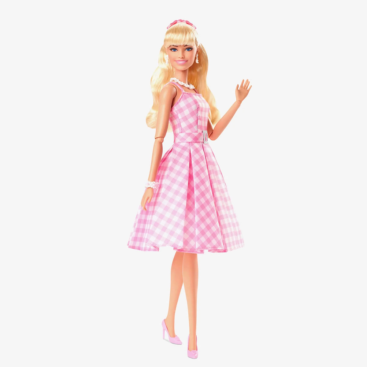 Lalka Barbie w różowej sukience w kratkę, machająca. Blond włosy, różowe buty i akcesoria. Białe tło.