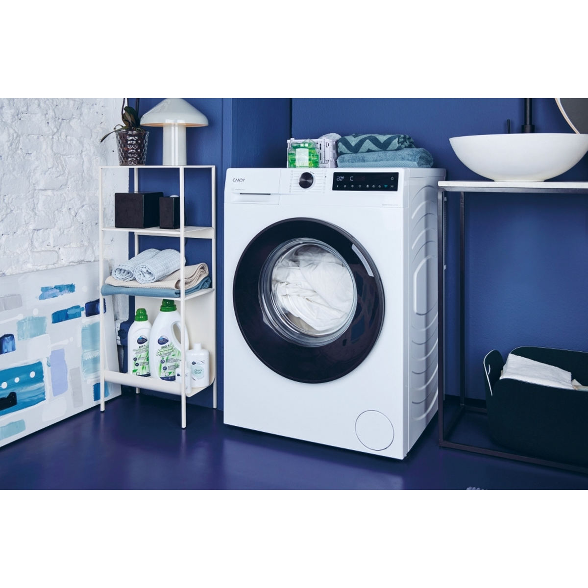 Lavadora con ropa blanca, estante con detergentes y pared azul al fondo.