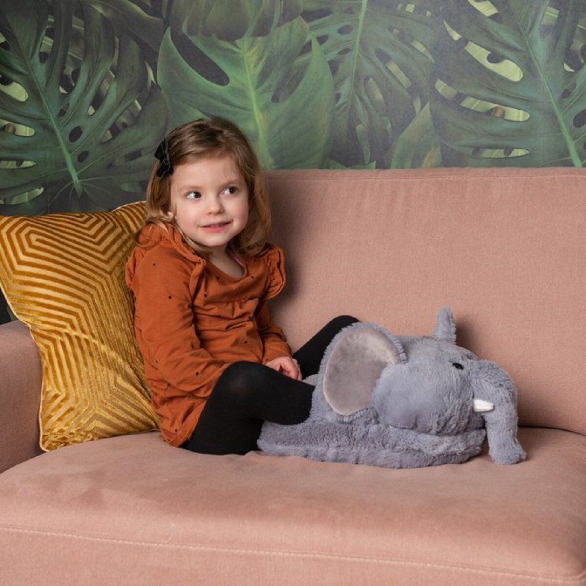 Una niña sentada en un sofá rosa con un elefante de peluche. Viste un vestido naranja y leggings negros. Una almohada amarilla a la izquierda.