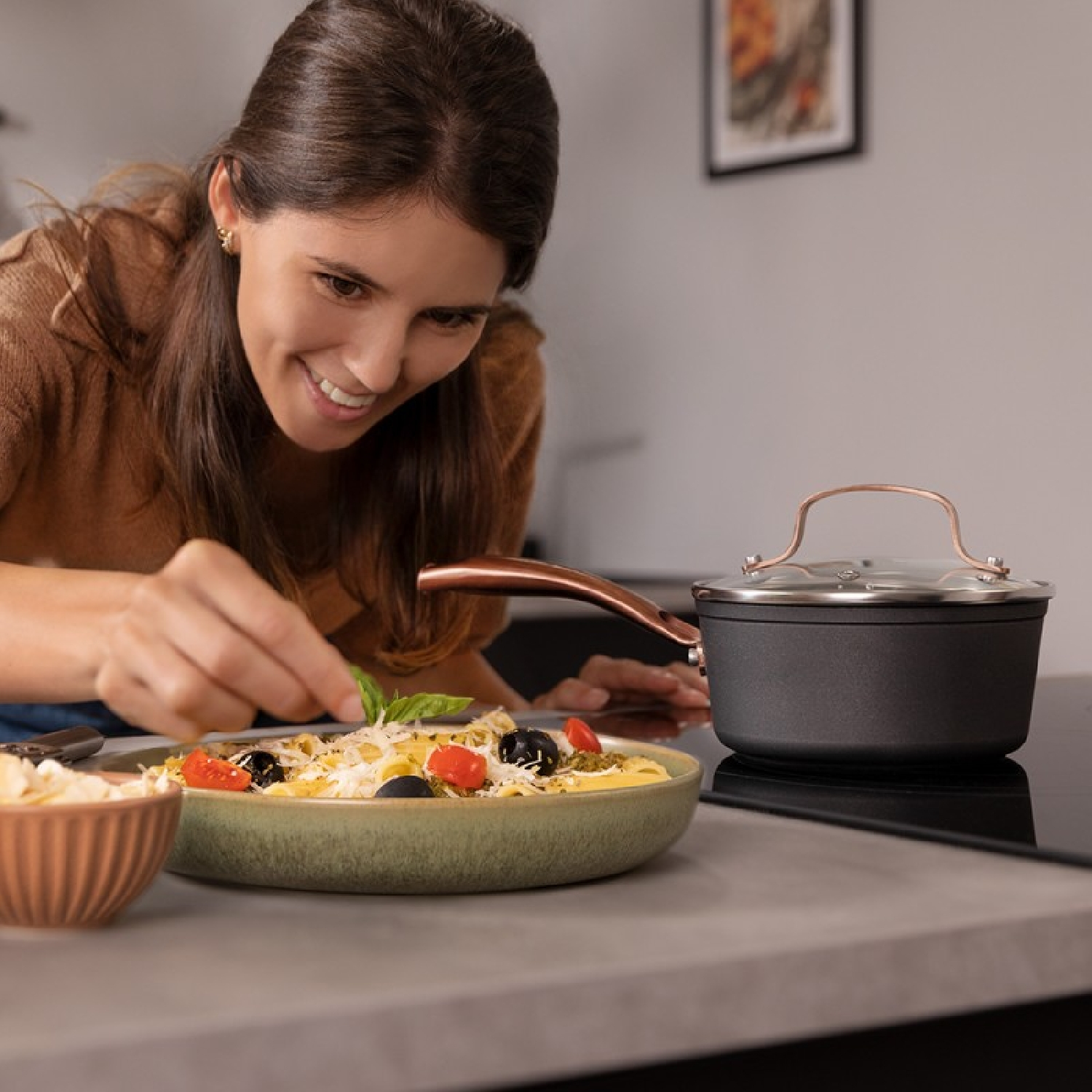 Mujer sonriendo en la cocina, agregando albahaca a pasta con tomates y aceitunas en un tazón.