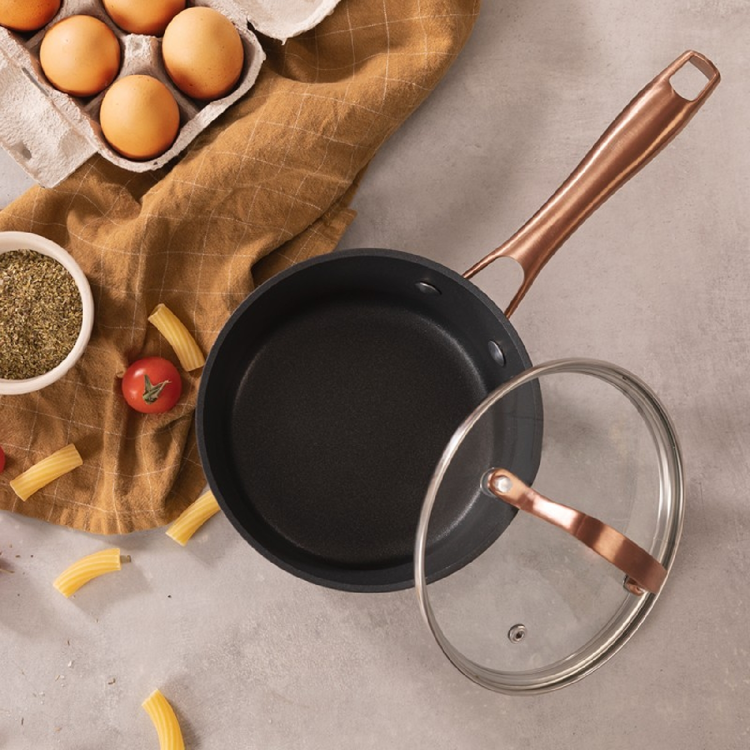 Sartén con mango de cobre, tapa quitada. Huevos, pasta y especias junto a la sartén.