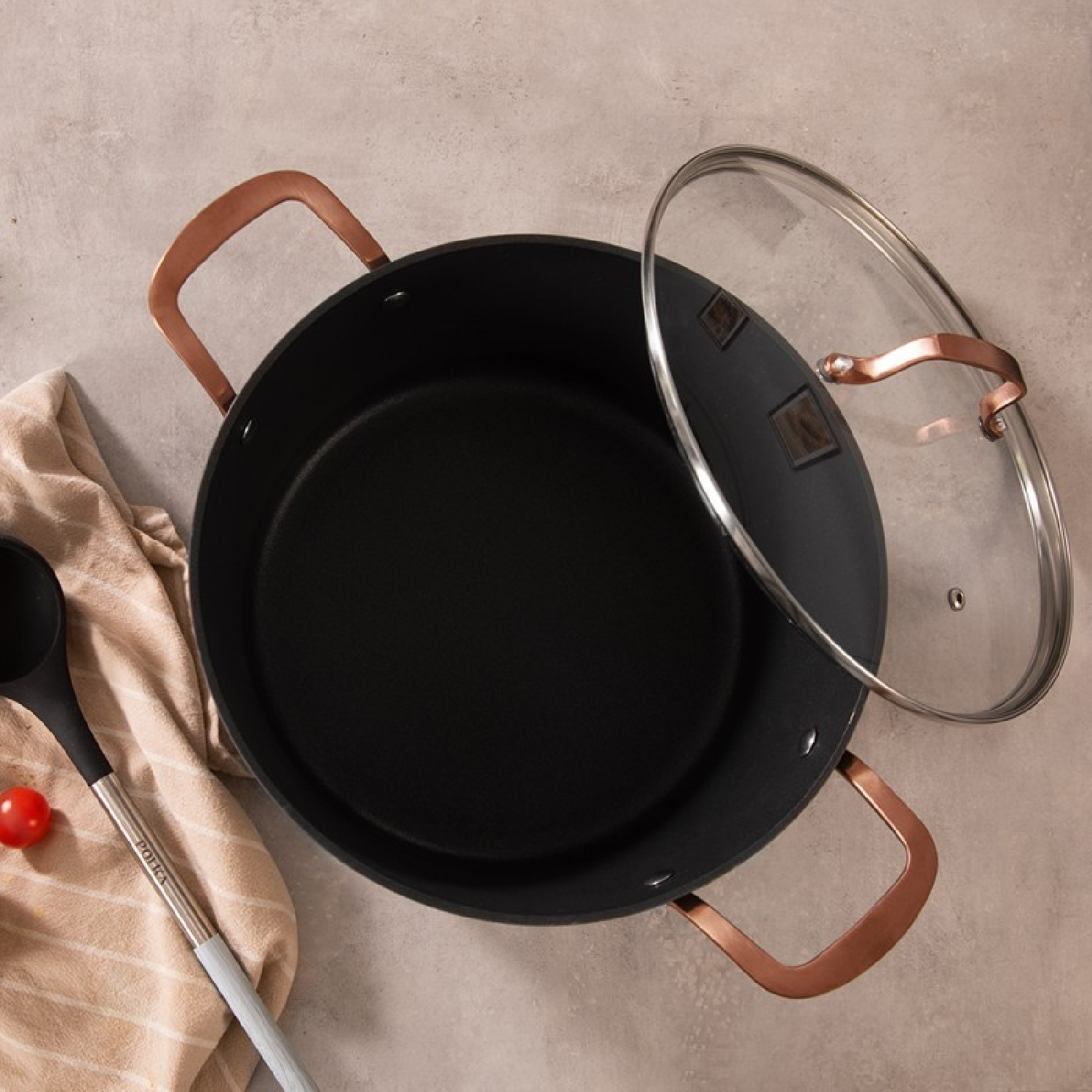 Zwarte wok met koperen handvatten, glazen deksel en etenswaren op een tafel.