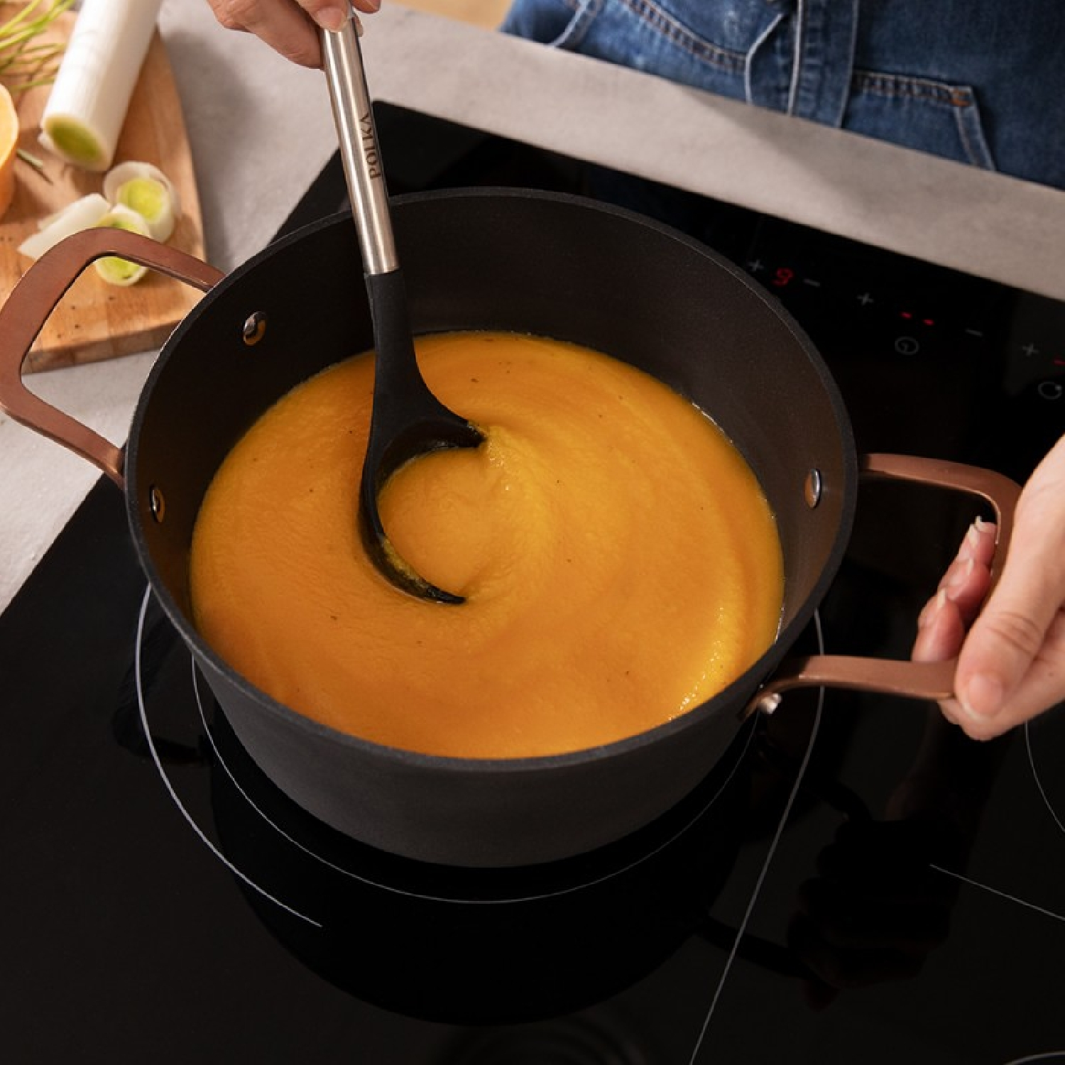 Una persona revuelve sopa de naranja en una olla negra con asas de cobre sobre una vitrocerámica negra.