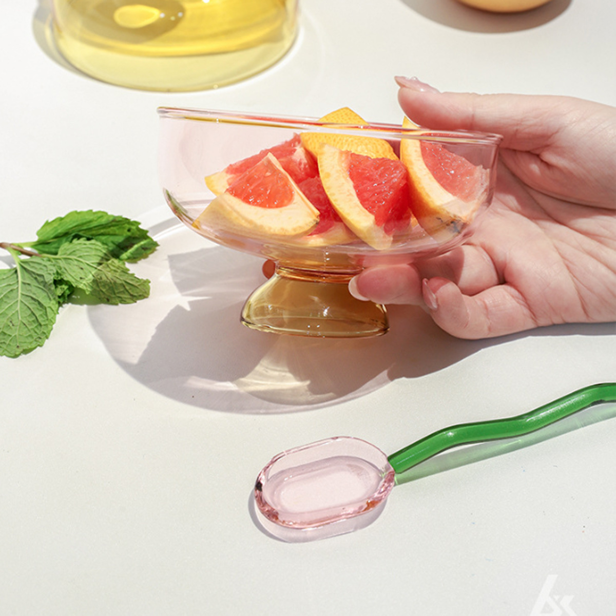 Eine Glasschale mit Grapefruit-Scheiben, ein Glaslöffel und eine Hand. Auf weißer Oberfläche, mit grünen Minzblättern.