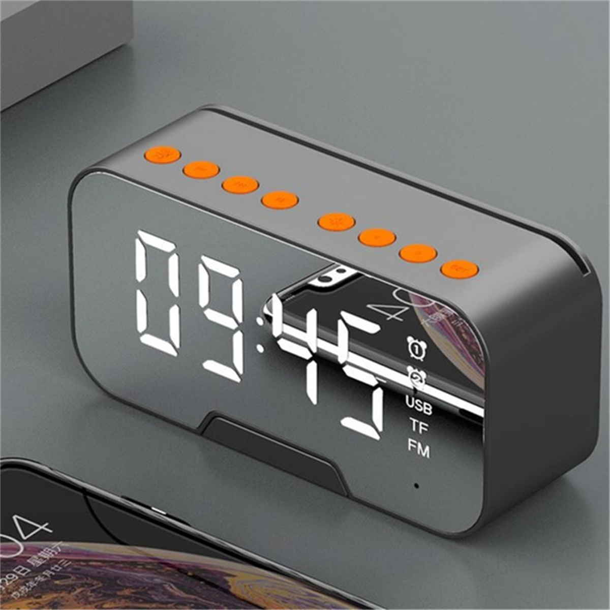 Ein digitaler Wecker, grau mit orangefarbenen Knöpfen, zeigt die Uhrzeit und spiegelt ein Telefon wider.