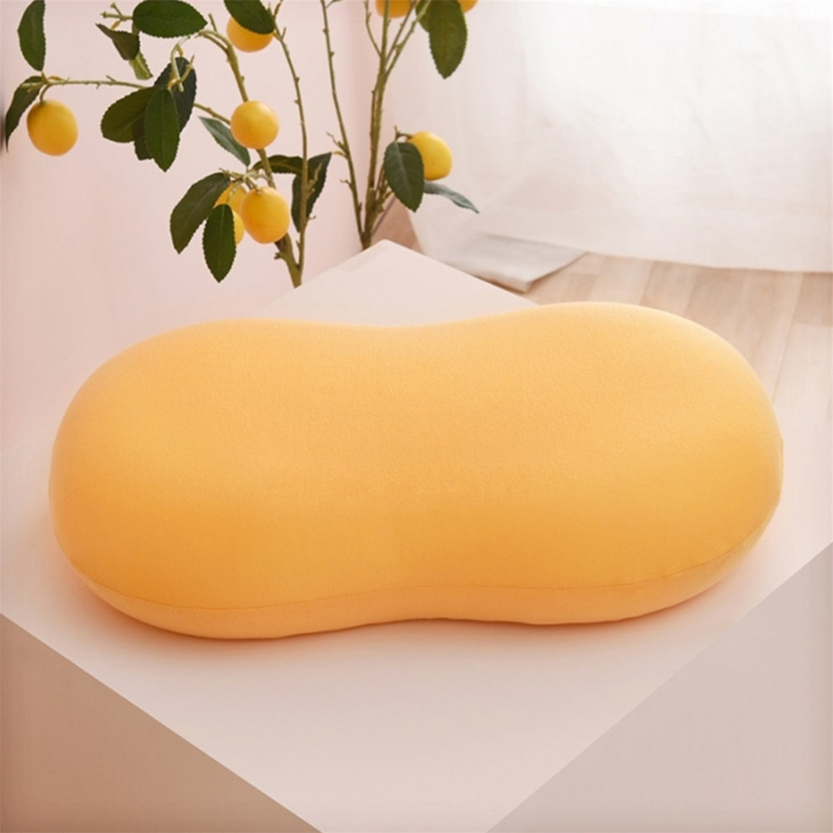 Ein gelbes Kissen in Erdnussform auf weißer Oberfläche. Dahinter eine Pflanze mit Zitronen.