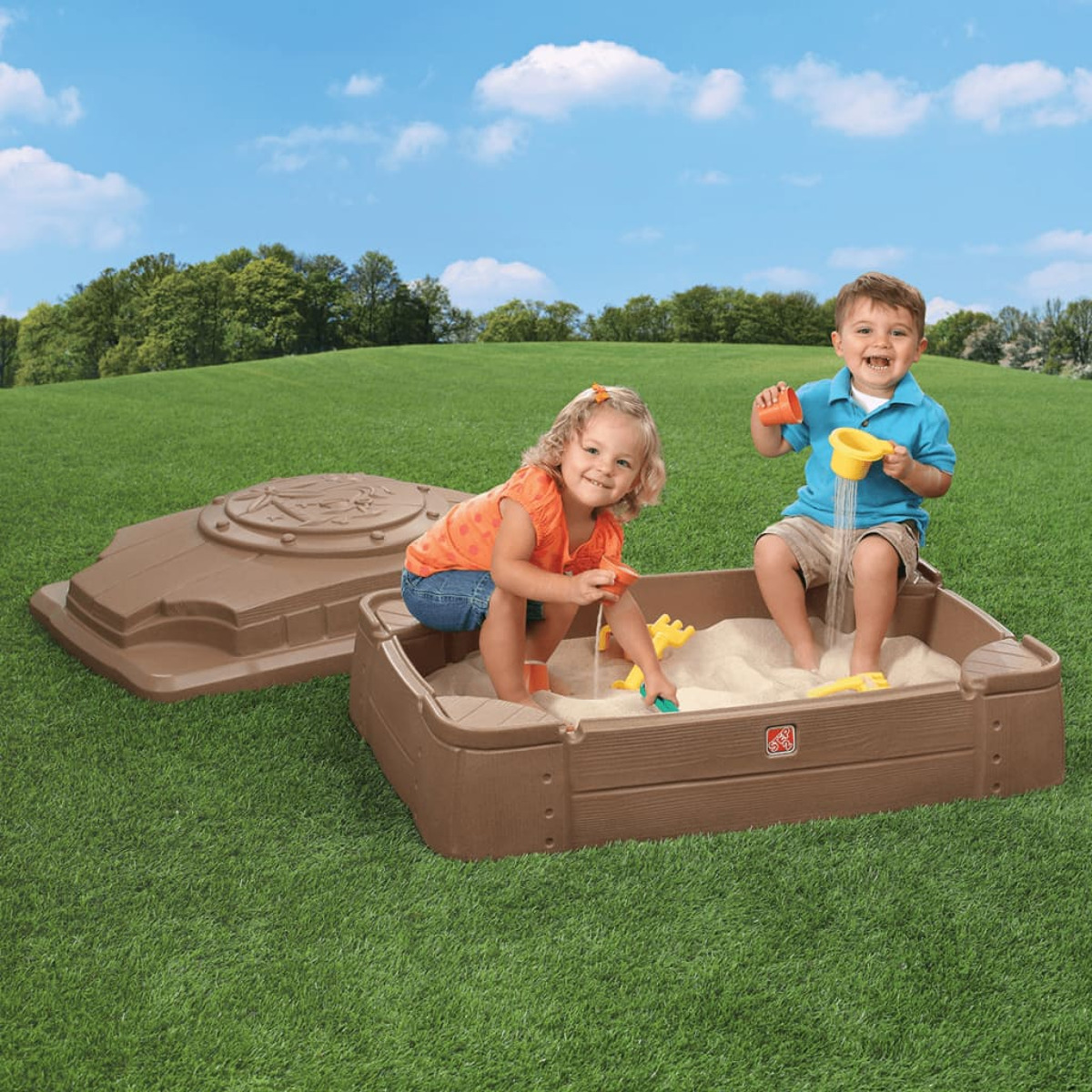 Zwei Kinder spielen in einem Sandkasten. Eines schüttet Sand mit einem gelben Becher aus. Das andere spielt mit einem Spielzeug. Brauner Sandkasten auf grünem Gras.