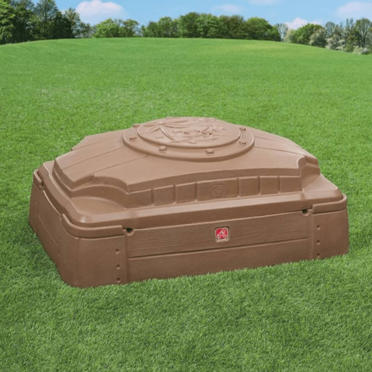 Ein brauner Sandkasten mit Deckel steht auf grünem Gras. Bäume und blauer Himmel sind im Hintergrund.