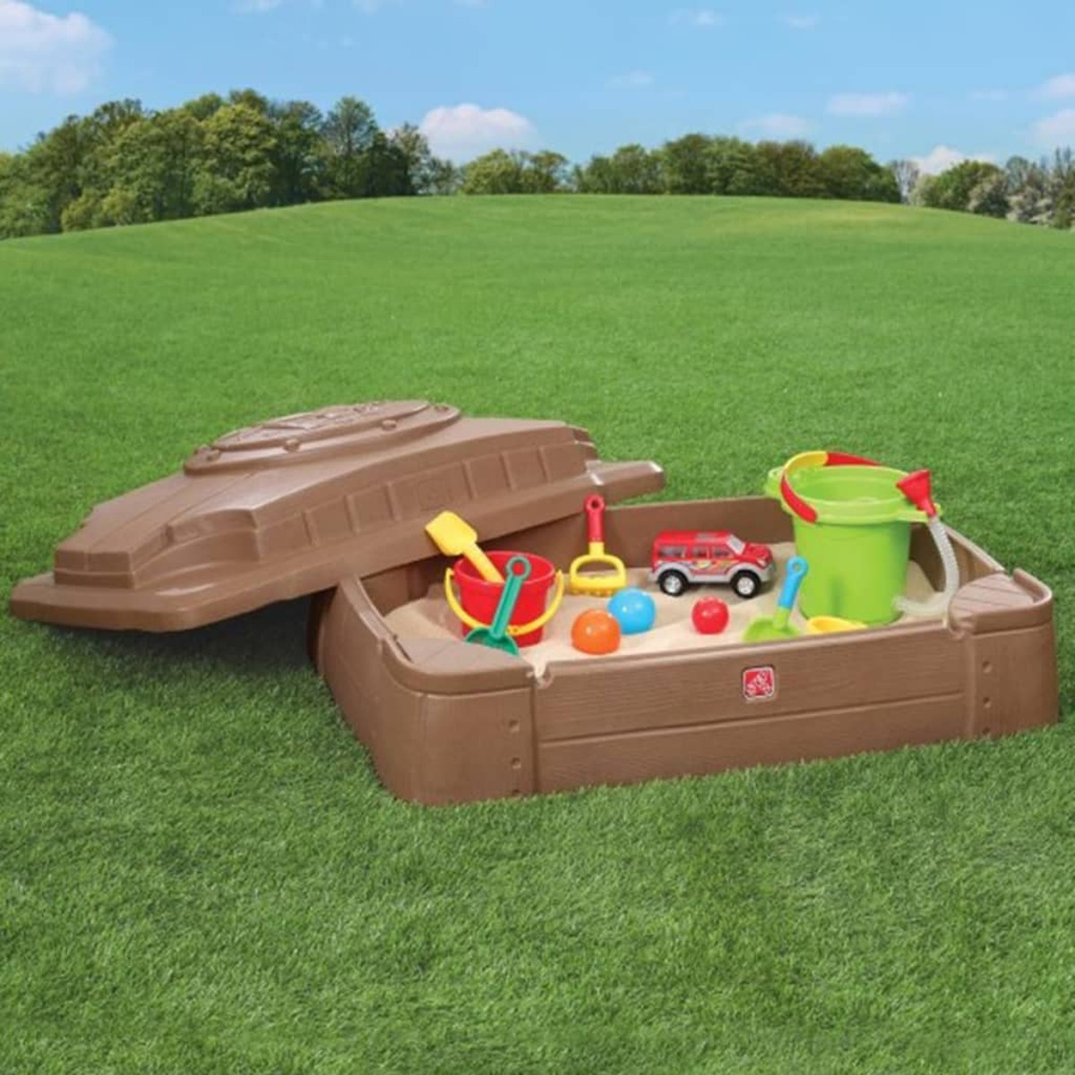 Ein brauner Sandkasten mit Spielzeug. Es hat einen Deckel. Spielzeug umfasst ein rotes Auto, Eimer, Schaufeln und Bälle. Grünes Gras und blauer Himmel im Hintergrund.