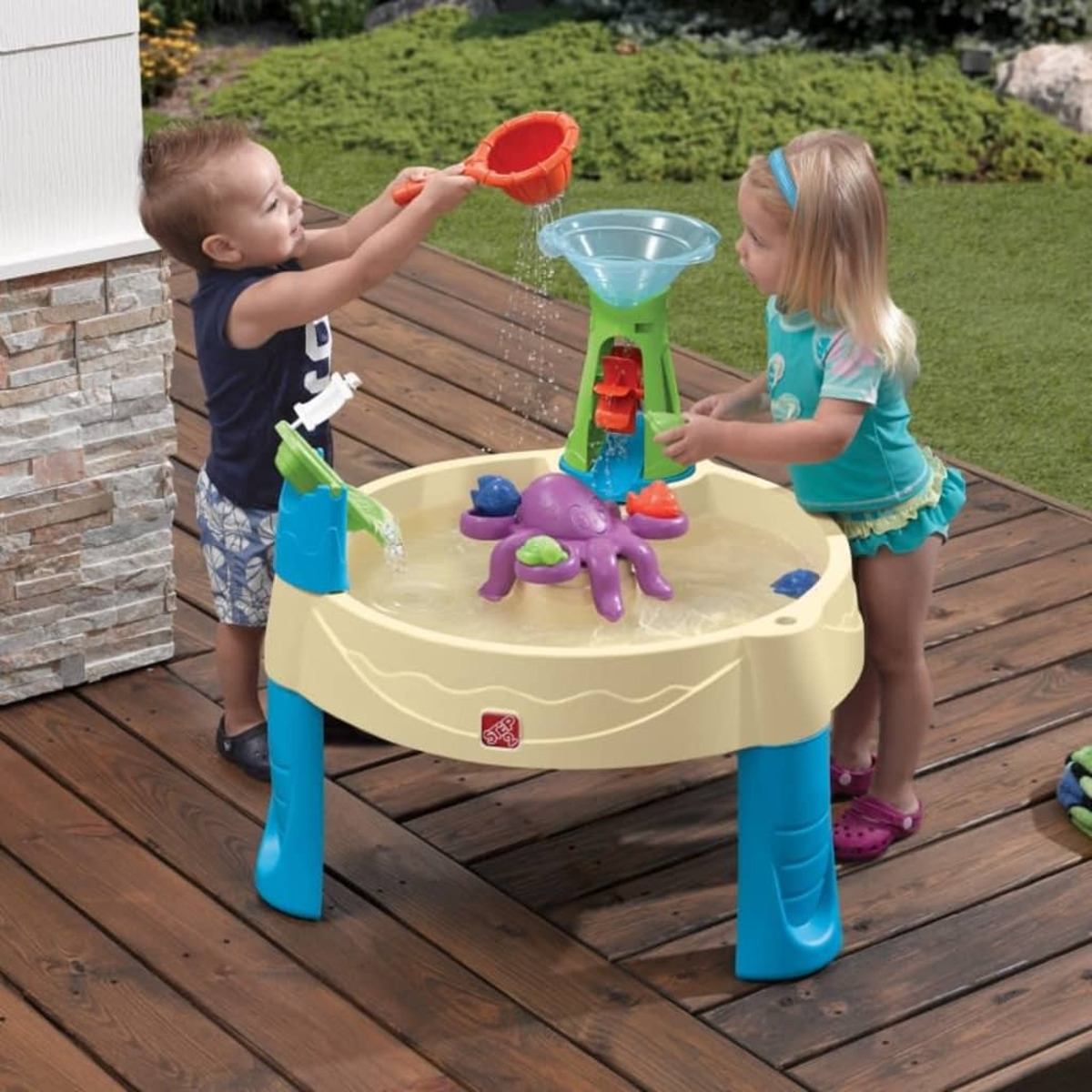 Zwei kleine Kinder spielen im Freien mit einem Wassertisch, sonniger Tag.