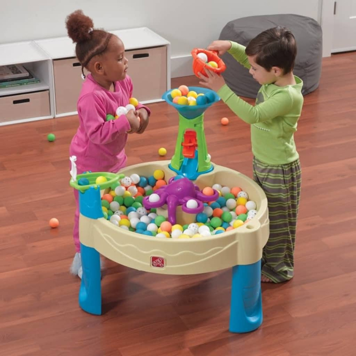 Zwei Kinder spielen mit bunten Bällen an einem Spieltisch. Beiger Tisch, blaue Beine.