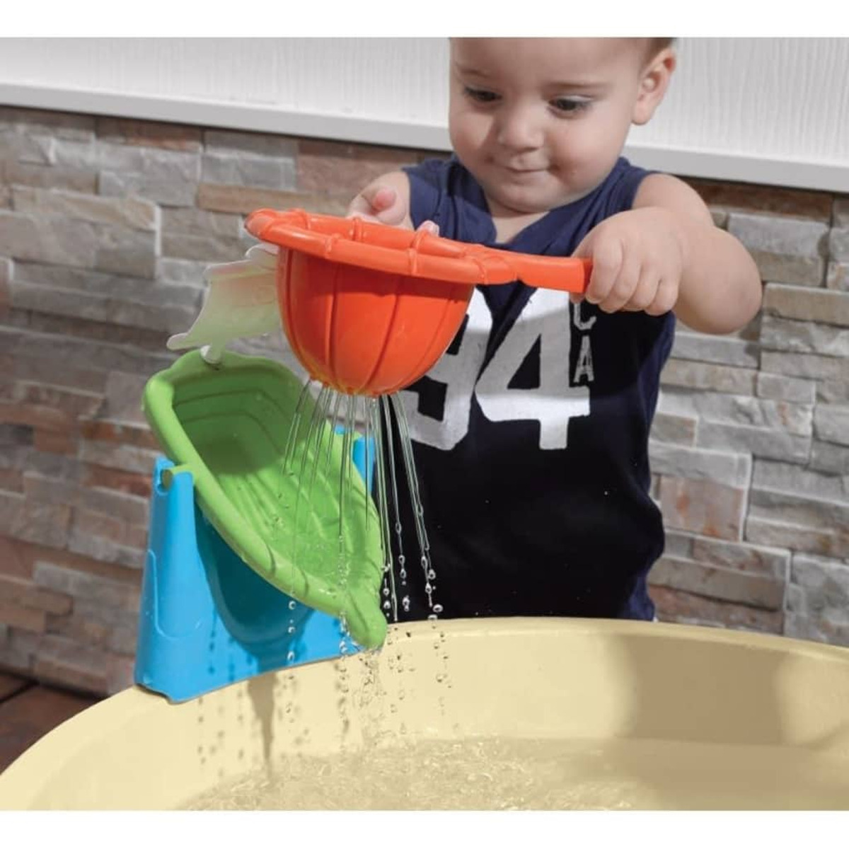 Ein Baby spielt mit einem Wasserspielzeug. Das Spielzeug gießt Wasser in eine gelbe Wanne. Hintergrund: Ziegelmauer.
