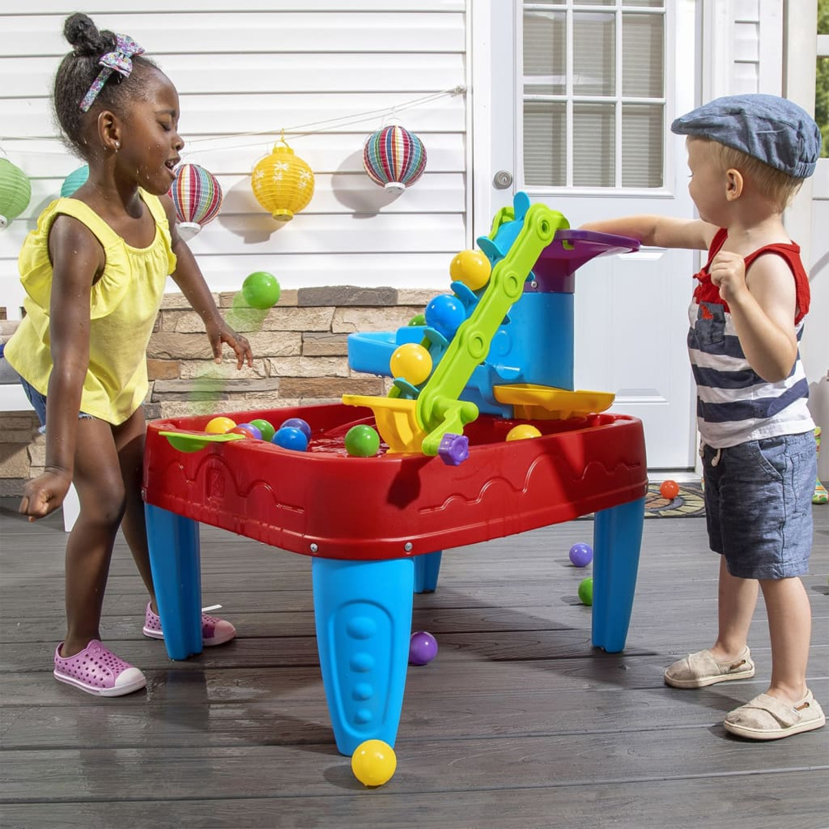 Zwei Kinder spielen mit einem Wassertisch, mit Bällen und Wasserfall, auf einer Terrasse.