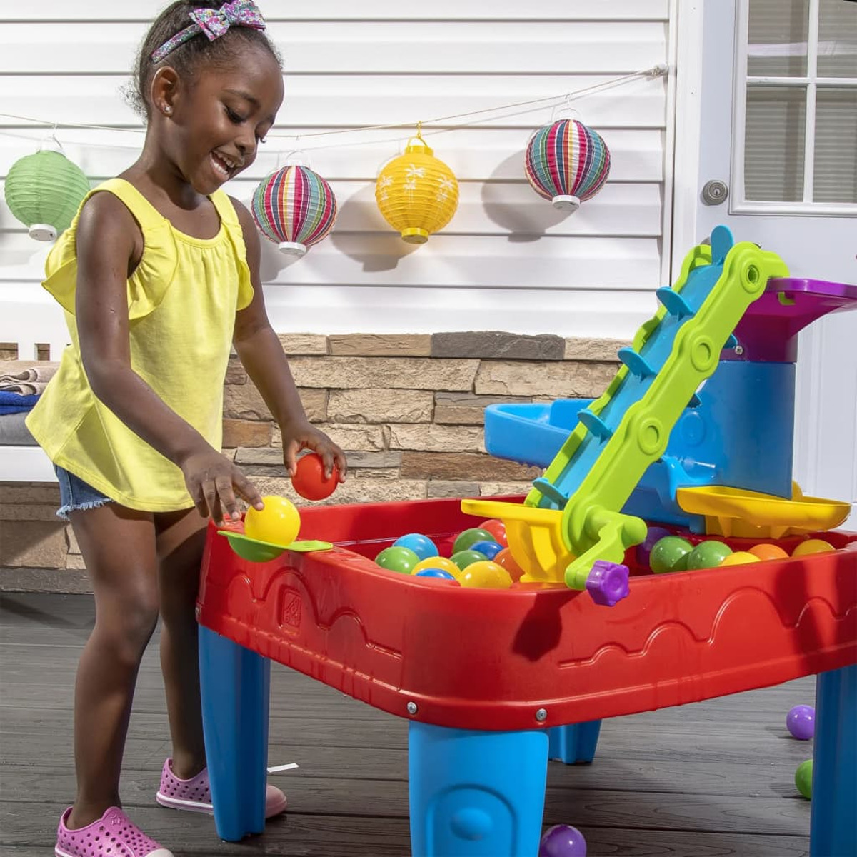 Ein Mädchen spielt mit einem Wassertisch, benutzt einen Schöpfer und Bälle. Bunte Laternen hängen im Hintergrund.