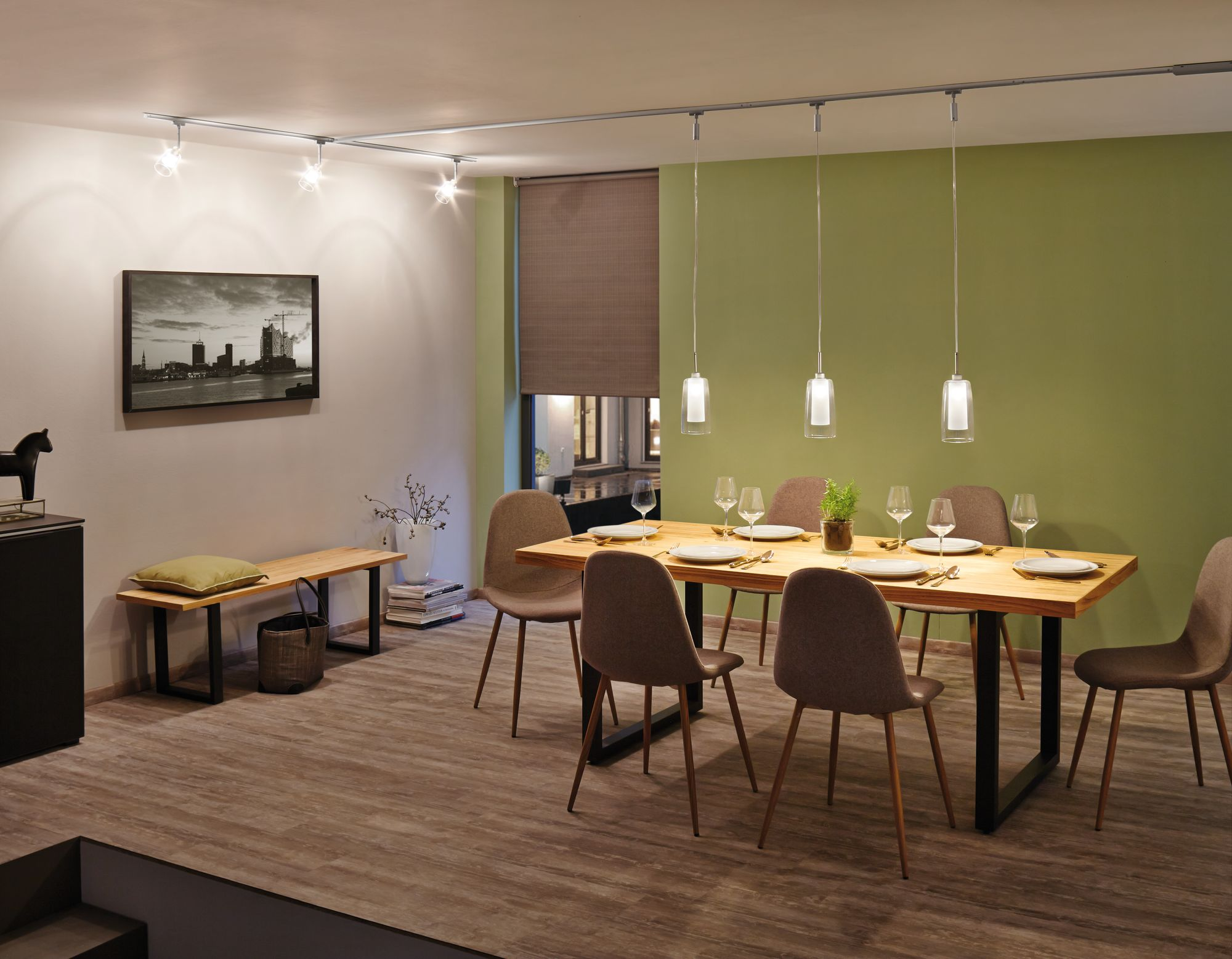 Esszimmer mit Holztisch, Stühlen und Pendelleuchten. Ein Bild und eine Bank sind an der Wand.