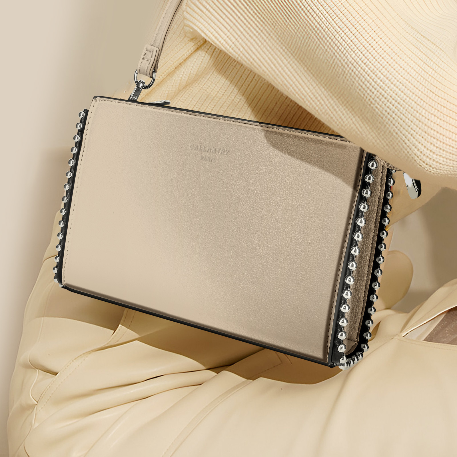 Beige Handtasche mit silbernen Nieten, gehalten von einer Person in cremefarbenem Pullover und Jacke.