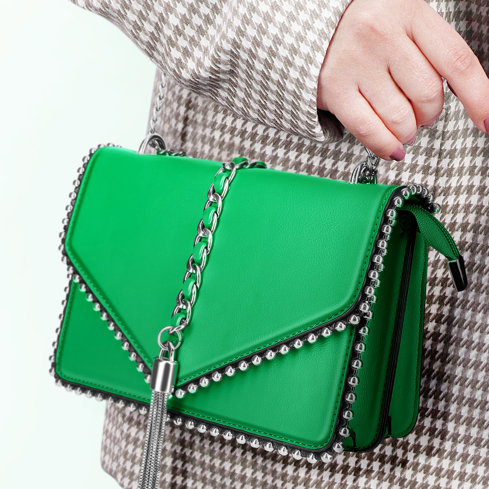 Grüne Handtasche mit silbernen Nieten und einem Kettenträger, gehalten von einer Person in einem gemusterten Mantel.