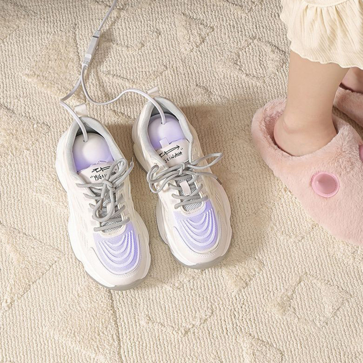 Scarpe bianche con luci e un cavo di alimentazione. Sono su un tappeto, accanto a una pantofola rosa.