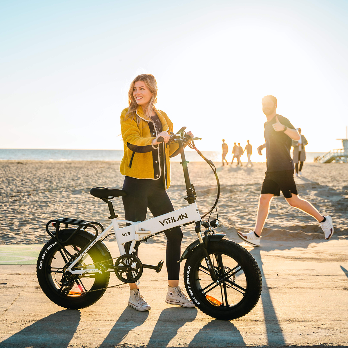 Kobieta z białym rowerem elektrycznym V3 na plaży, w tle inna osoba biegnie.