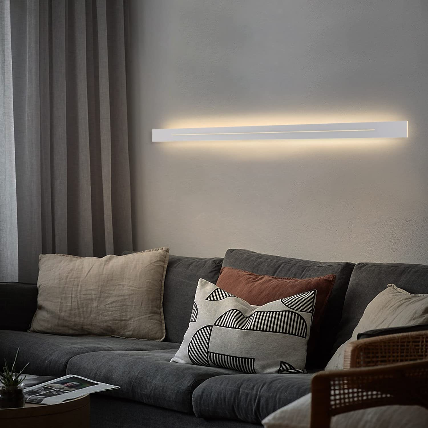 Un lungo apparecchio di illuminazione bianco montato a parete sopra un divano grigio. I cuscini poggiano sul divano.