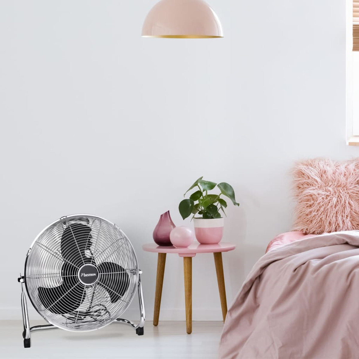 Ventilatore cromato sul pavimento in una stanza rosa. Tavolo rosa con pianta e cuscino rosa.