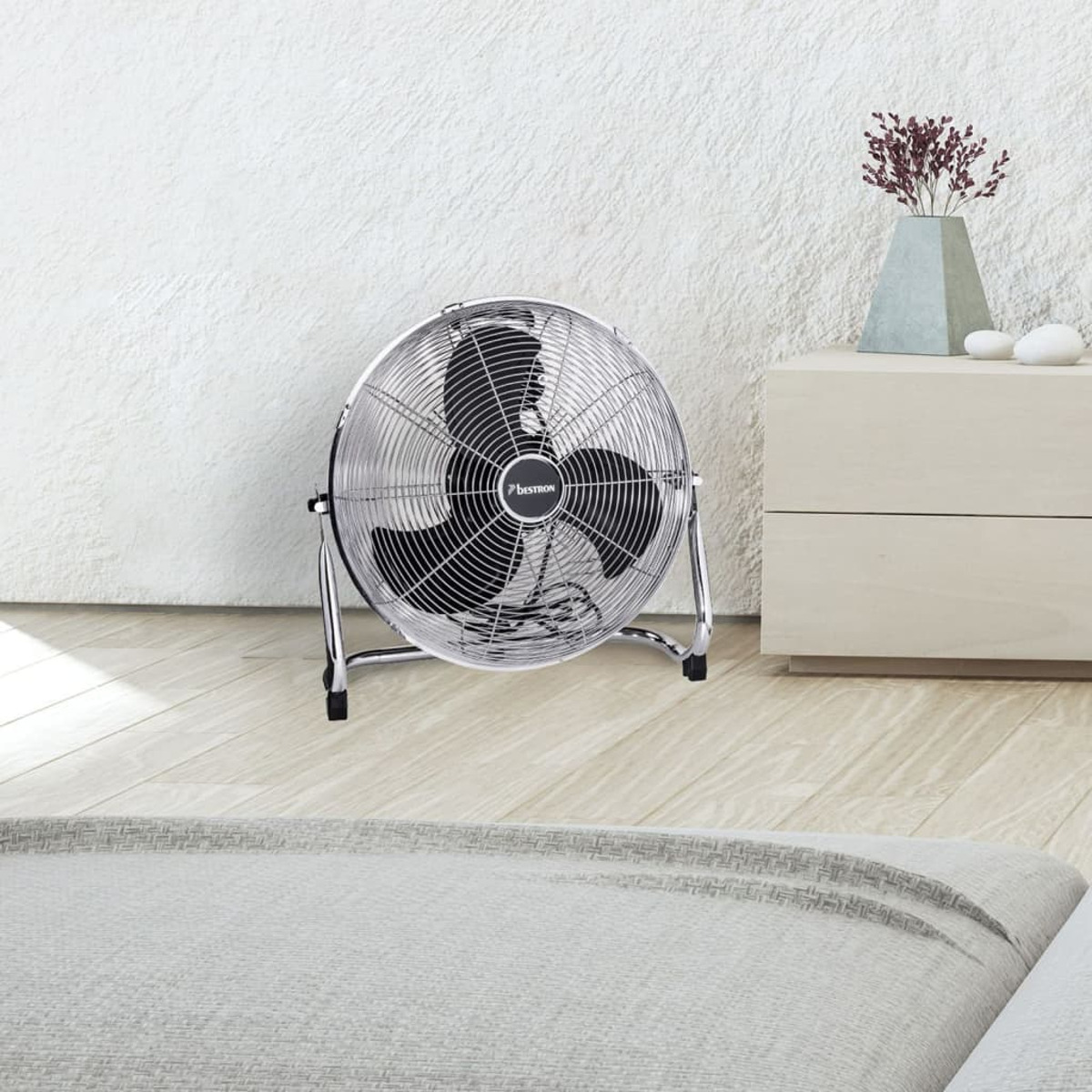 Un ventilatore argentato in una stanza. Si trova sul pavimento. Un vaso e un comodino sono sullo sfondo.