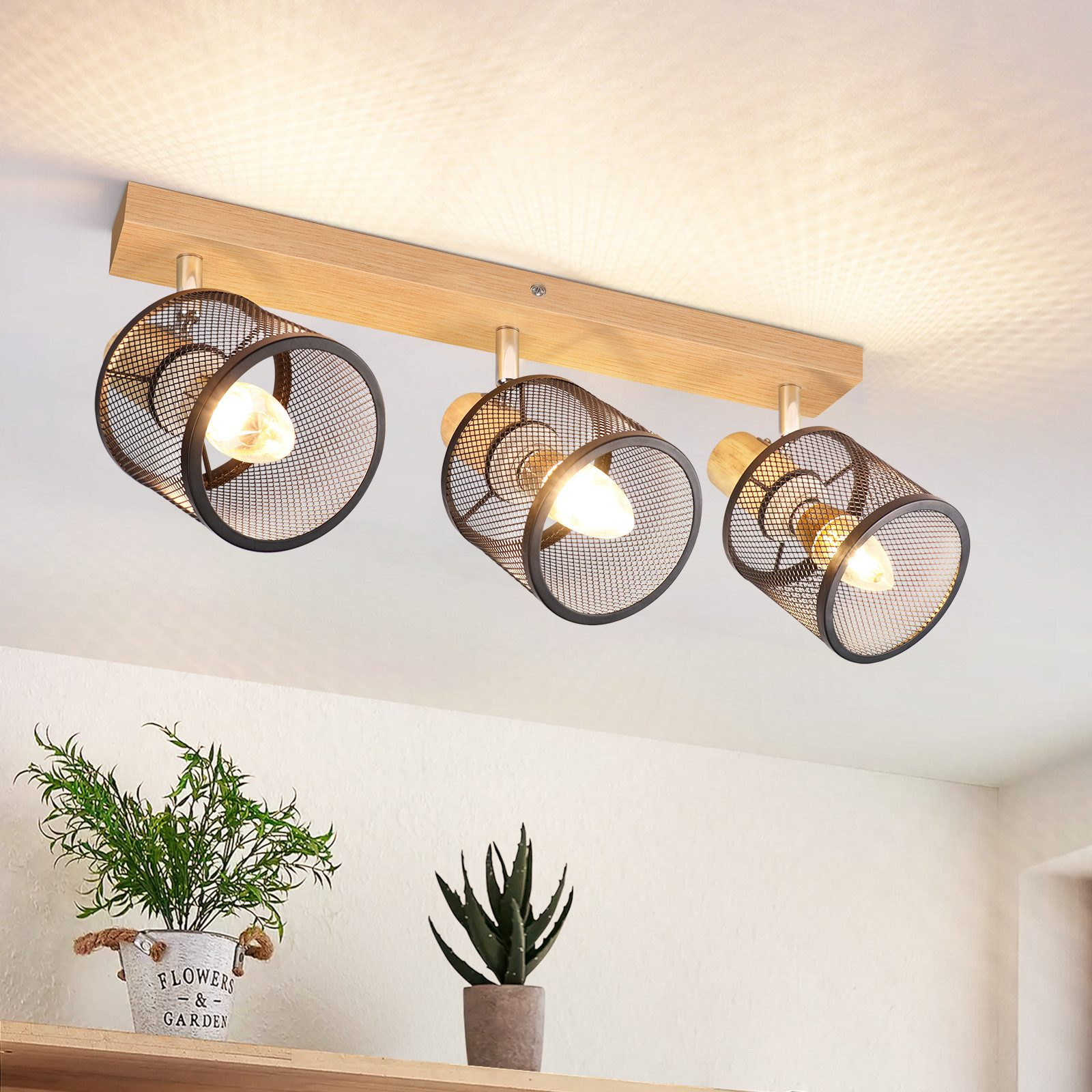 Lampy sufitowe z kloszami z siatki i drewnianą belką. Rośliny w doniczkach na pierwszym planie. Emitują ciepłe światło.