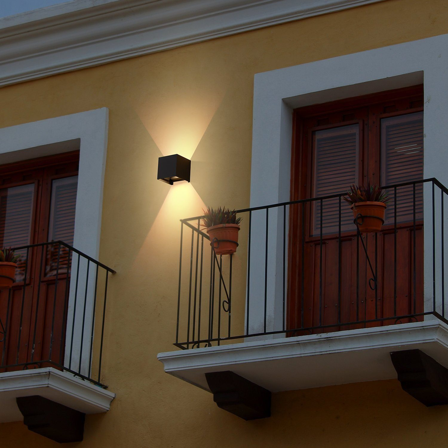 Widok zewnętrzny domu z czarną lampą ścienną na żółtej ścianie i balkonem.
