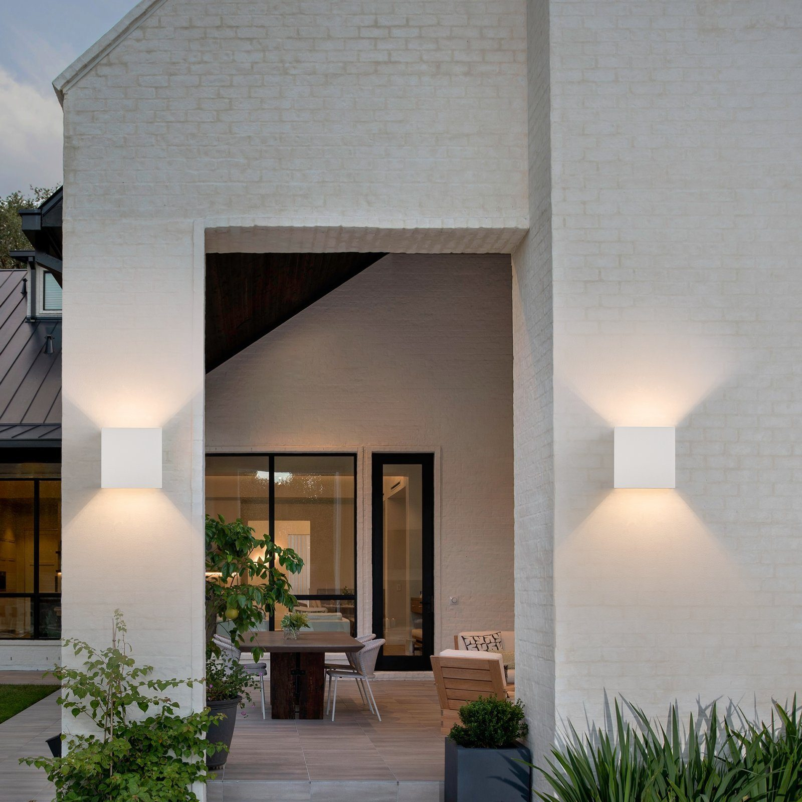 Ujęcie zewnętrzne wejścia do domu z kwadratowymi białymi lampami ściennymi. Dobrze oświetlone, z roślinami.