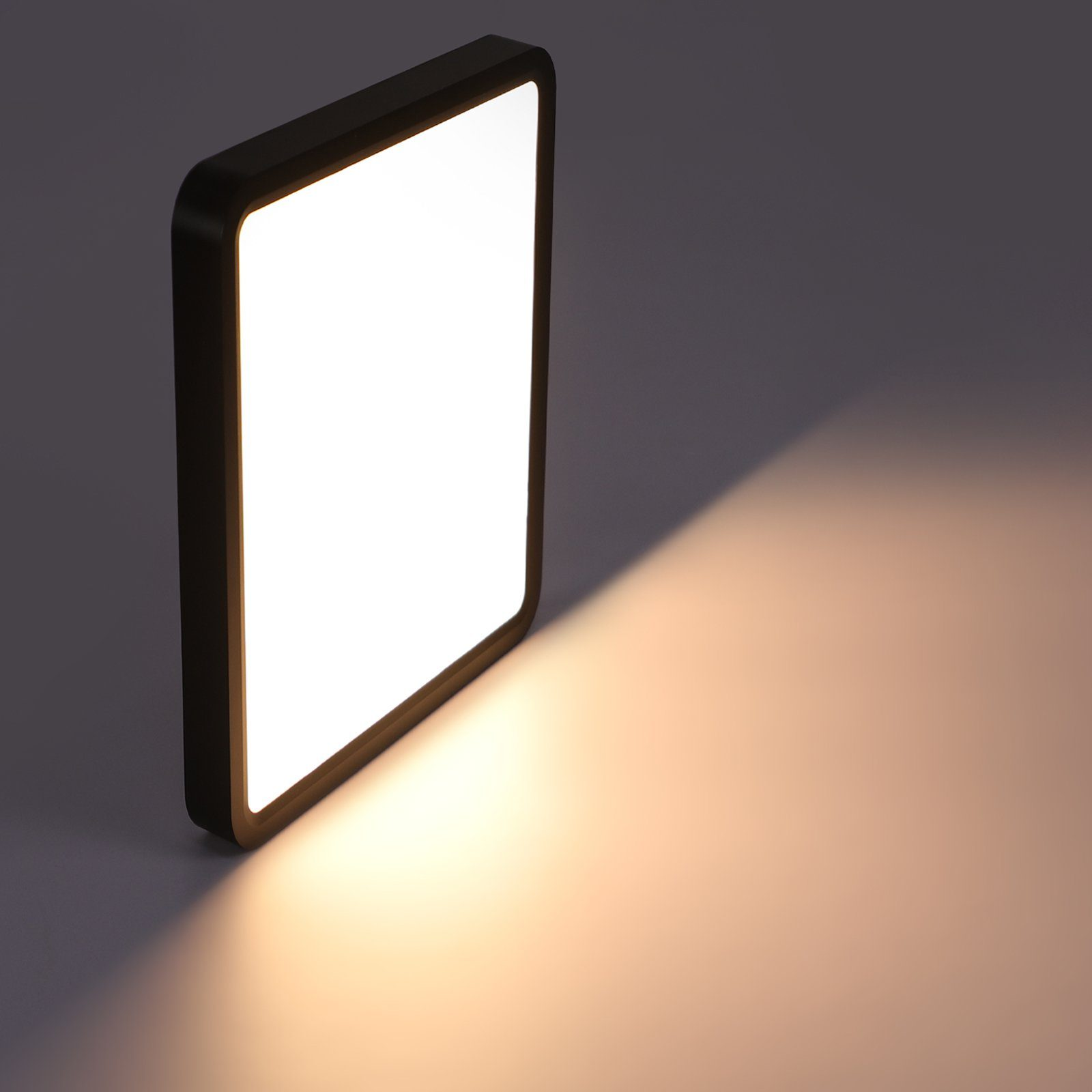 Kwadratowa, podświetlona lampa z czarną ramą rzuca ciepłe światło i cień na szarą powierzchnię.