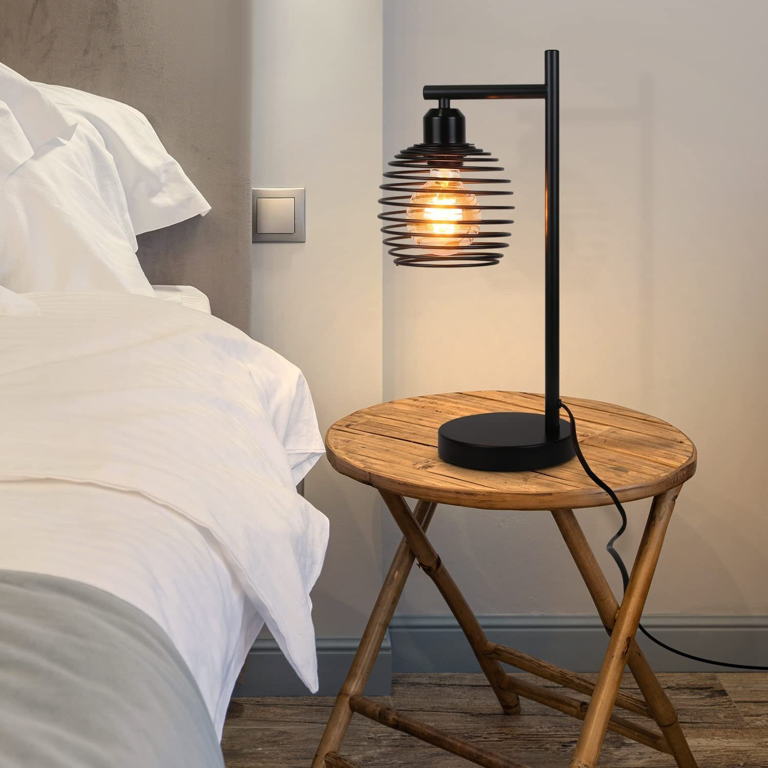 Czarna metalowa lampa stołowa z kloszem w kształcie spiralnej klatki i żarówką Edisona, na drewnianym stole.
