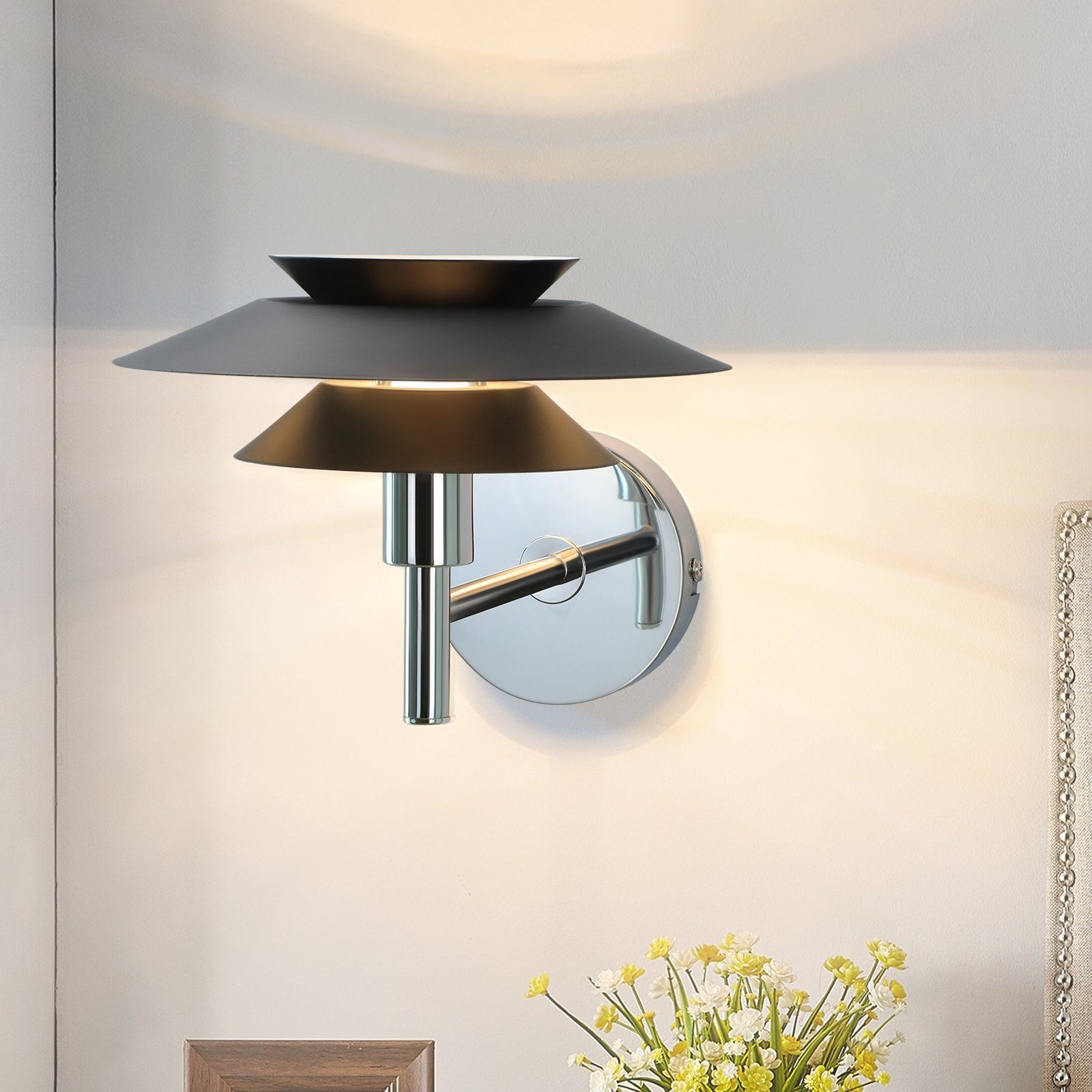 Czarno-srebrna lampa ścienna o warstwowym designie. Zamontowana na białej ścianie z kwiatami.