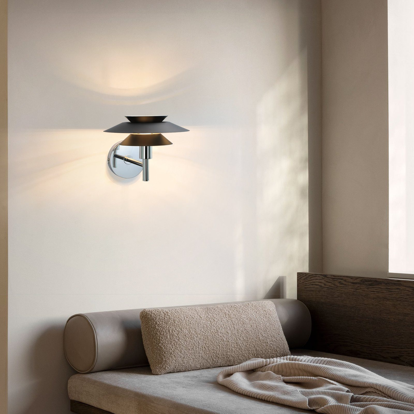 Nowoczesna lampa ścienna na beżowej ścianie, nad beżową sofą z poduszką i kocem.