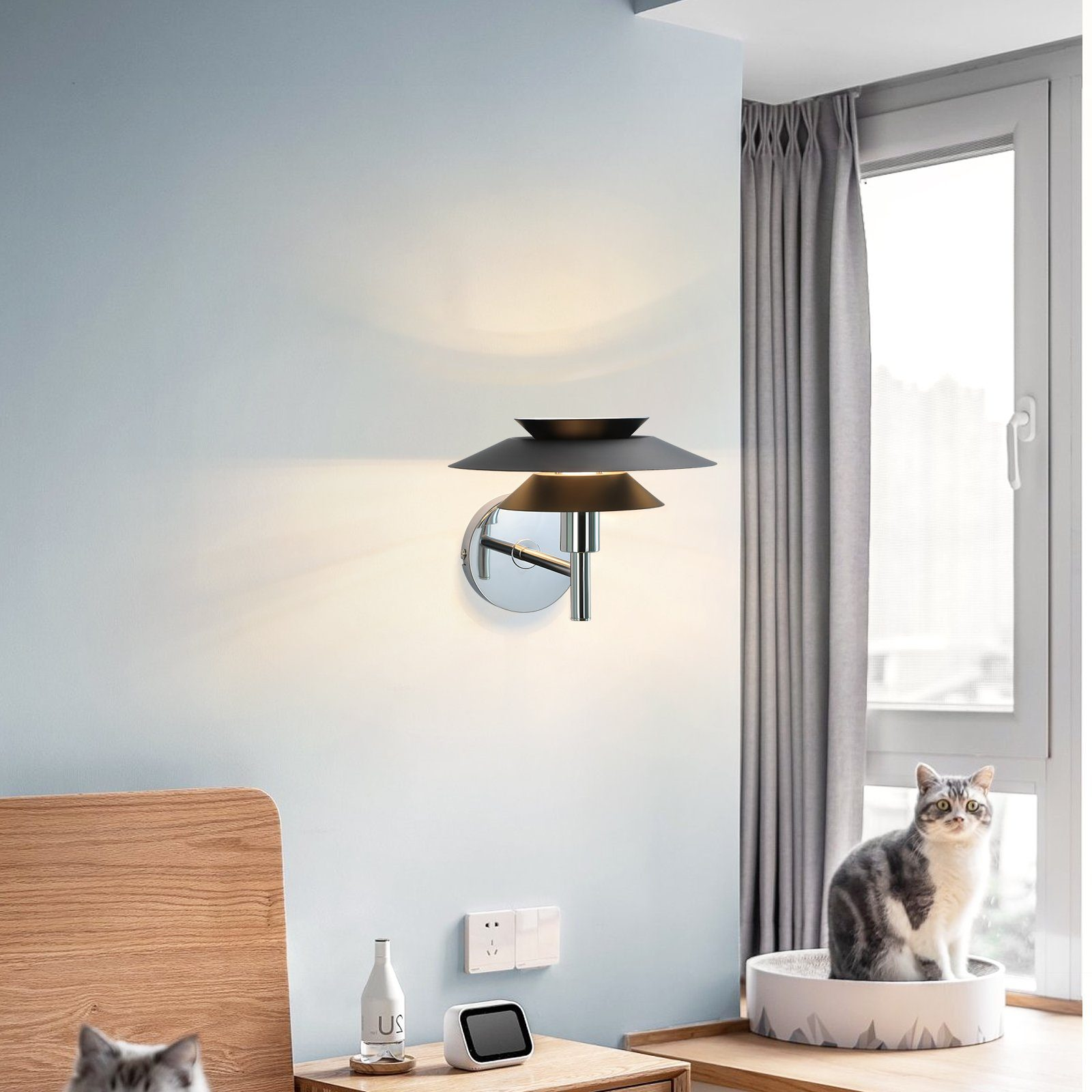 Nowoczesna lampa ścienna z trzema nałożonymi na siebie kloszami. Kot siedzi na posłaniu dla kota, obok okna.