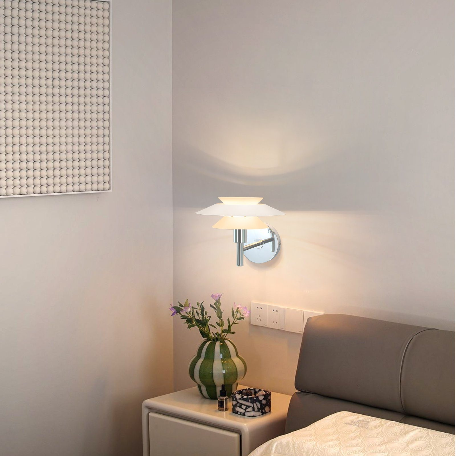 Lampa ścienna ze srebrną podstawą i białymi kloszami. Wazon z kwiatami stoi na stole.