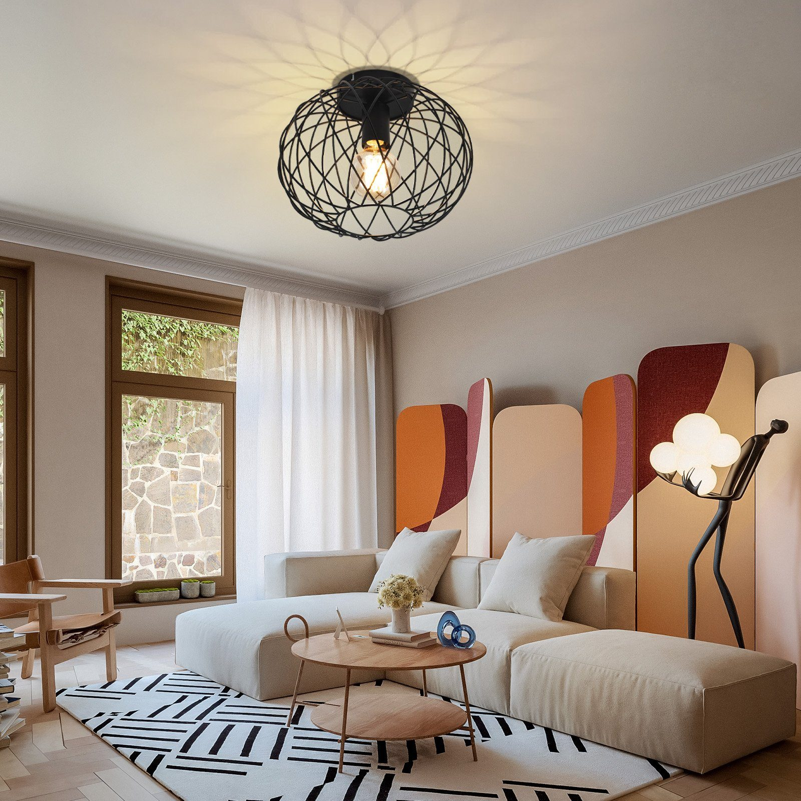 Czarna metalowa lampa sufitowa z drucianą ramą z żarówką. Na tle sofa, dywan i kolorowe panele.