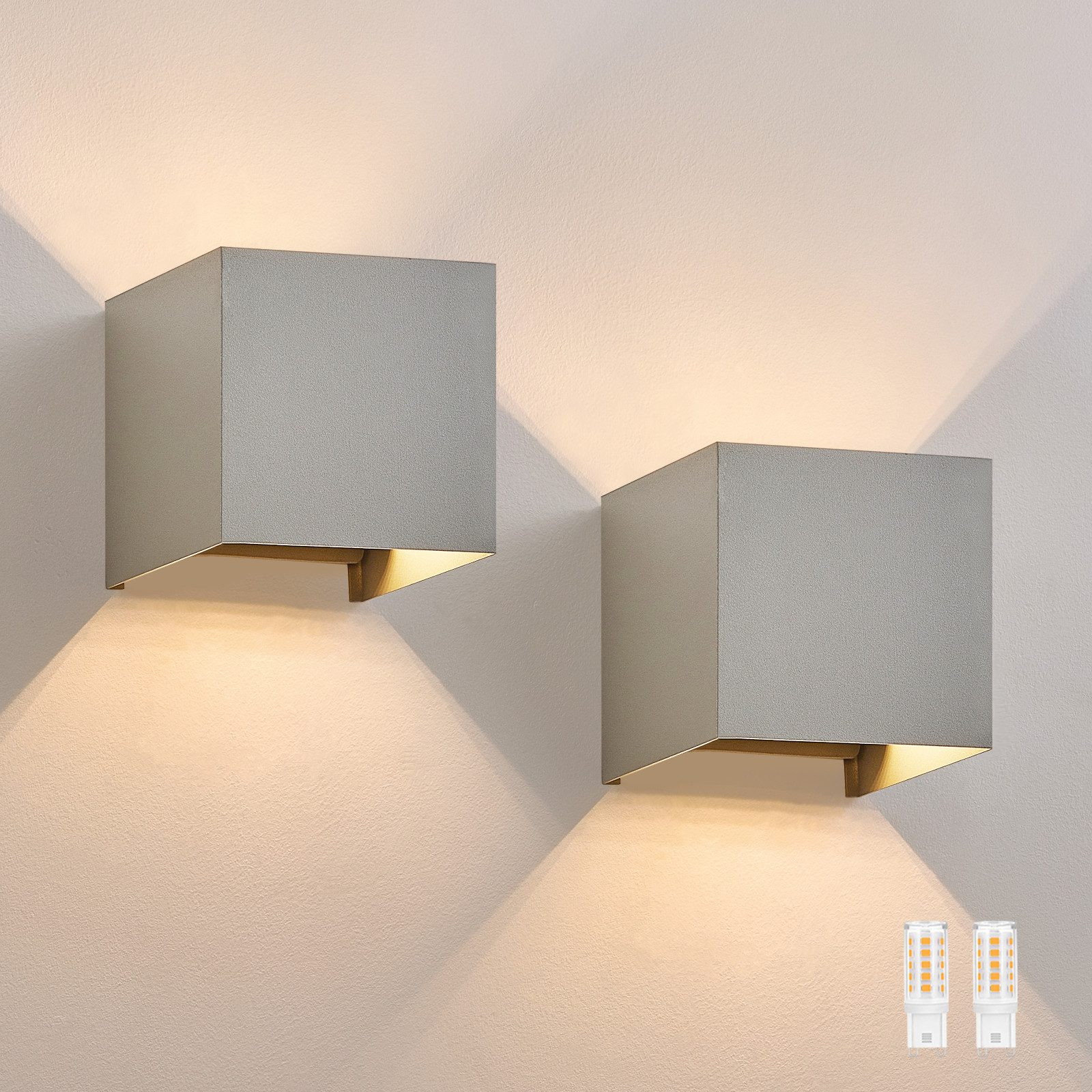 Dwie srebrne kwadratowe lampy ścienne z ciepłym blaskiem na jasnoszarej ścianie.