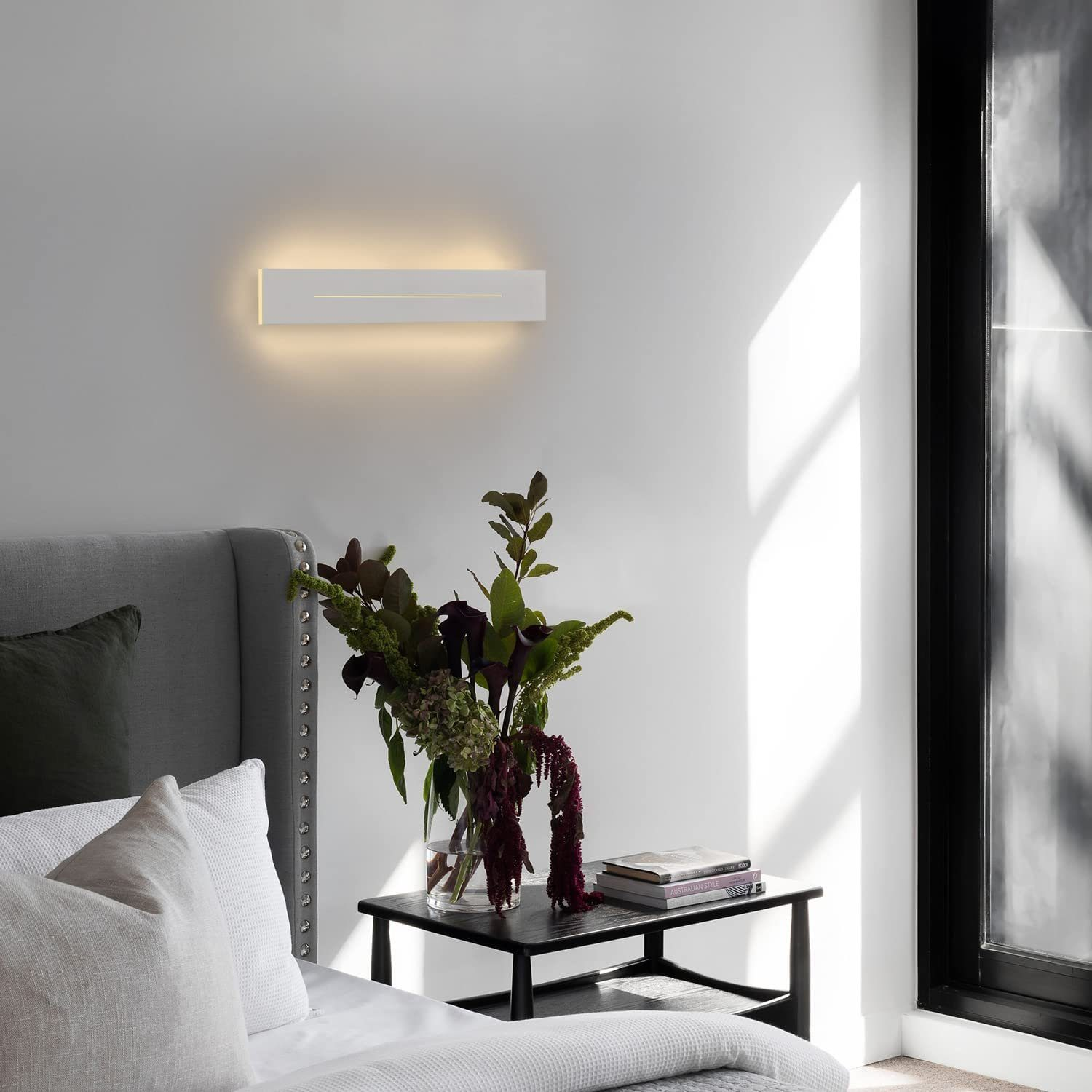 Nowoczesna sypialnia z białą lampą ścienną nad stołem z kwiatami, książkami i łóżkiem na pierwszym planie.